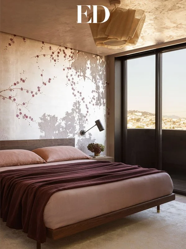 A bedroom with a wooden bed frame, pink pillows, and a dark red blanket, a white nightstand with a plant, a wall light, a textured wall with cherry blossom artwork, and a large window showing a city view.