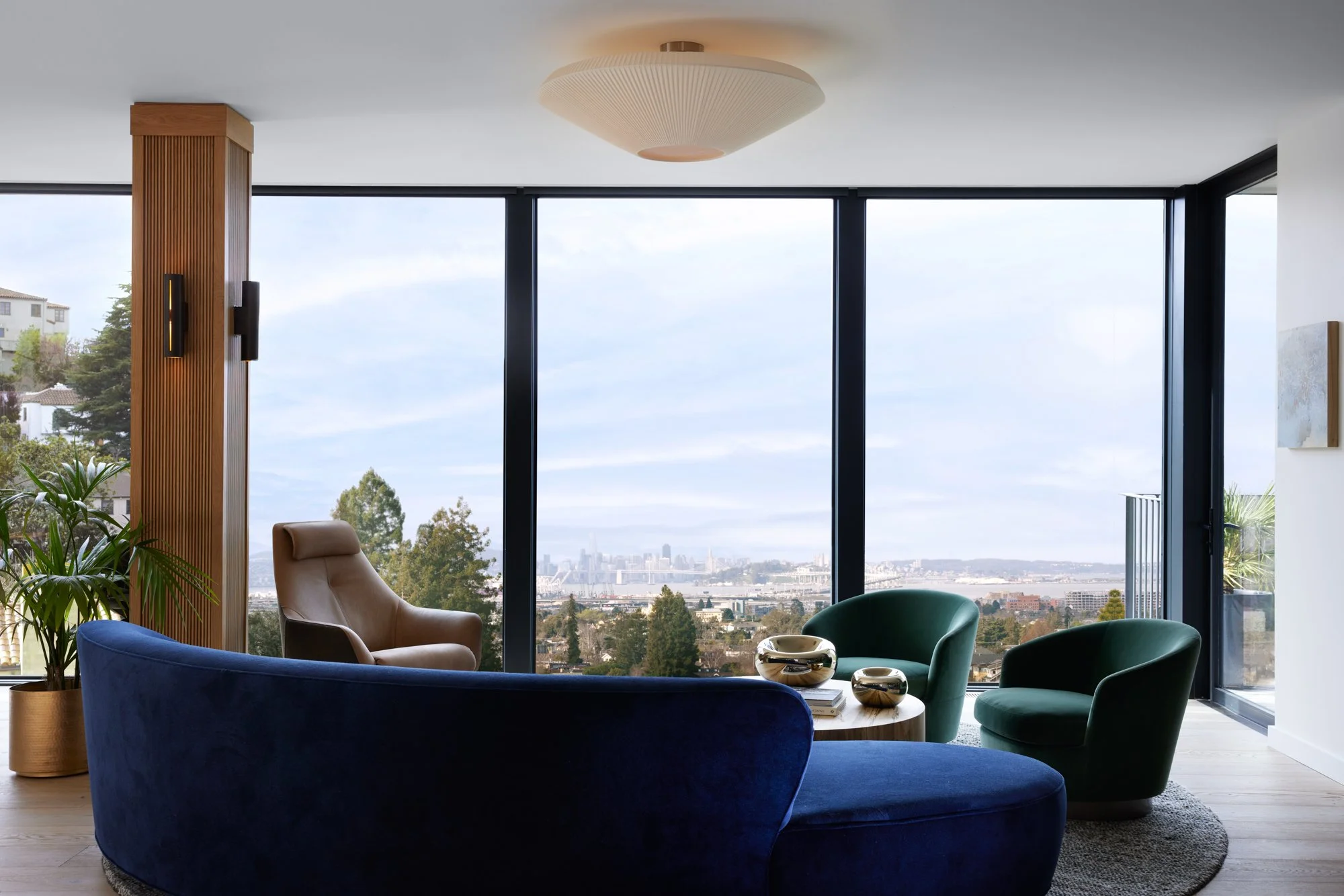 Modern living room with floor-to-ceiling windows overlooking the city, featuring colorful armchairs, a beige chair, a wooden coffee table, and decorative bowls, with plants and a textured ceiling light.