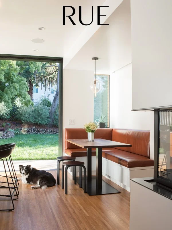 Modern dining area with a built-in booth with orange cushions, a rectangular table, a small black stool, a potted plant, and a view of a green yard with trees through glass doors. There is a black and white dog lying on the wooden floor.