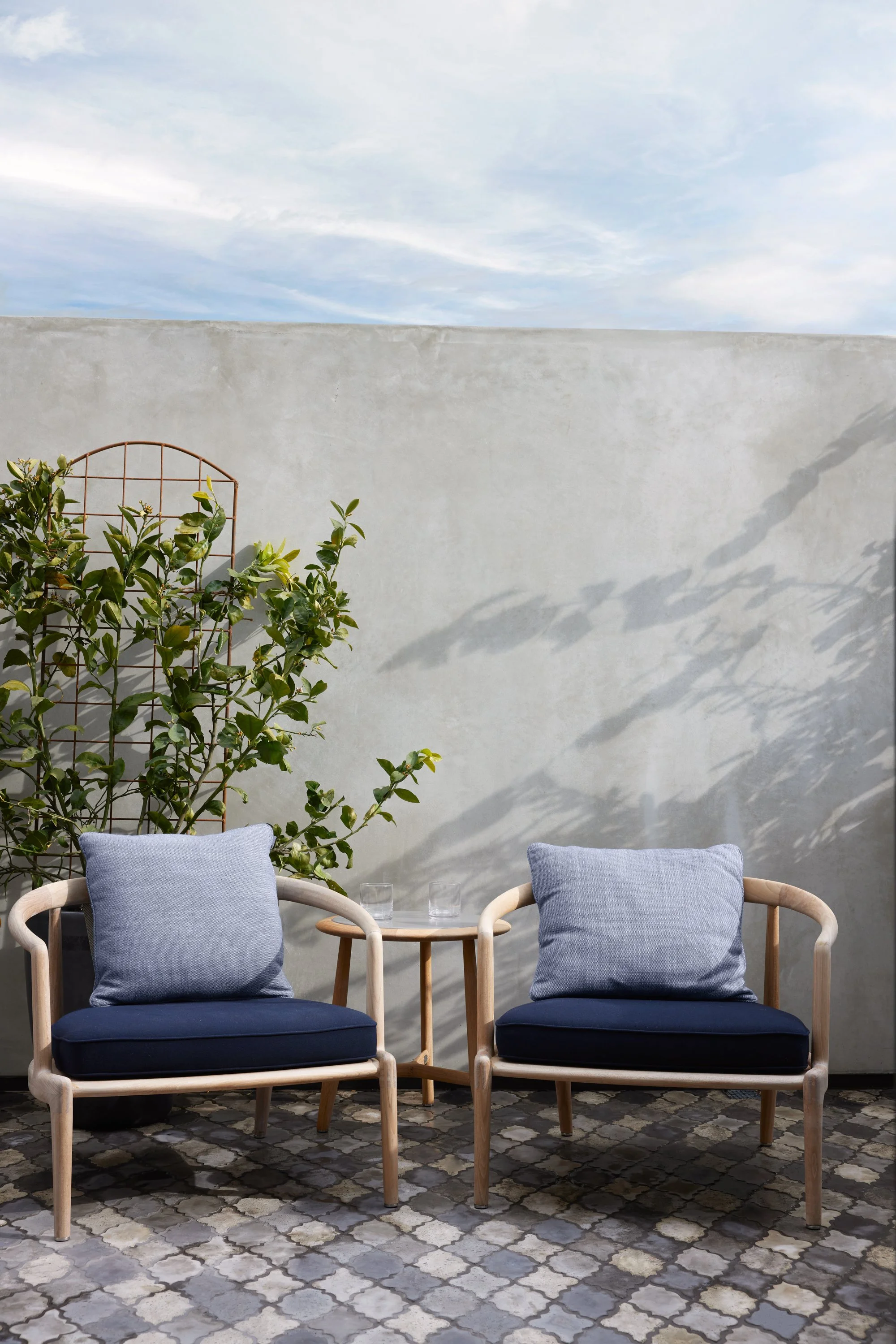 Two wooden chairs with light blue cushions and a small wooden table with two glasses on it, set against a plain gray wall with shadows, a potted plant, and a patterned tile floor.