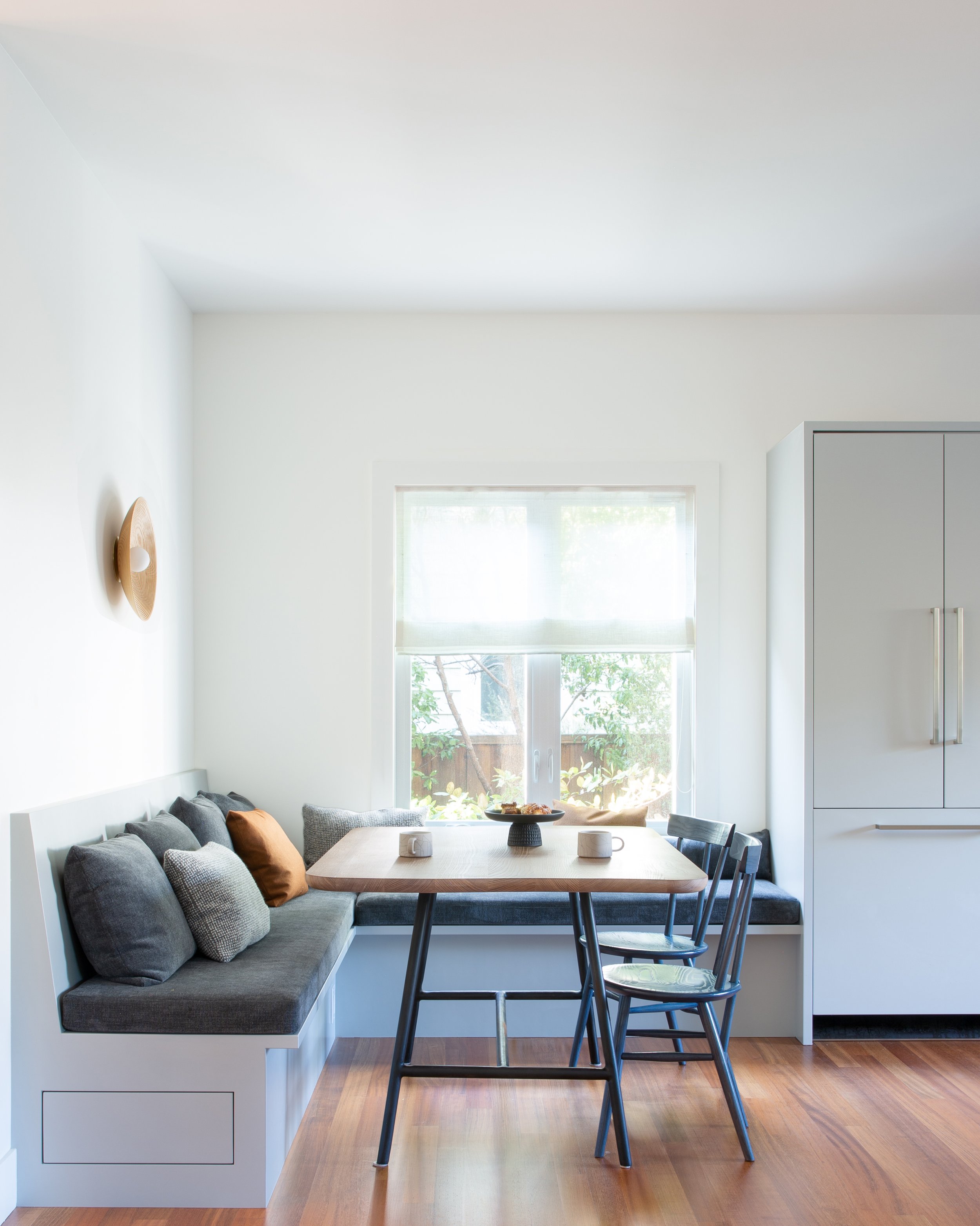 A bright dining area with a gray upholstered bench with gray, white, and rust-colored pillows, a wooden table, a black chair, and a window showing greenery outside.