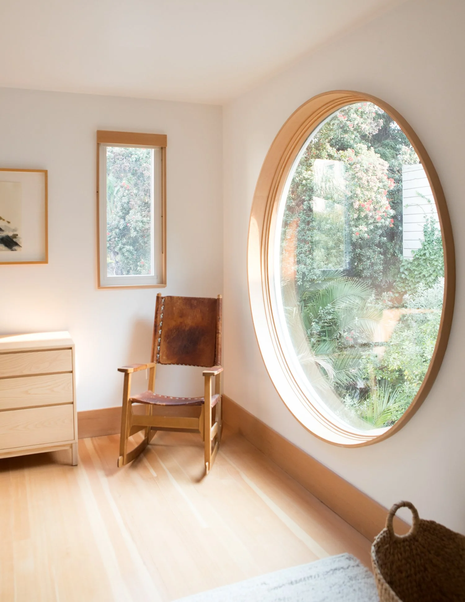 A cozy room with a large round window showing lush greenery outside. There is a wooden chair and a small wooden dresser in the corner, with a framed artwork on the wall.