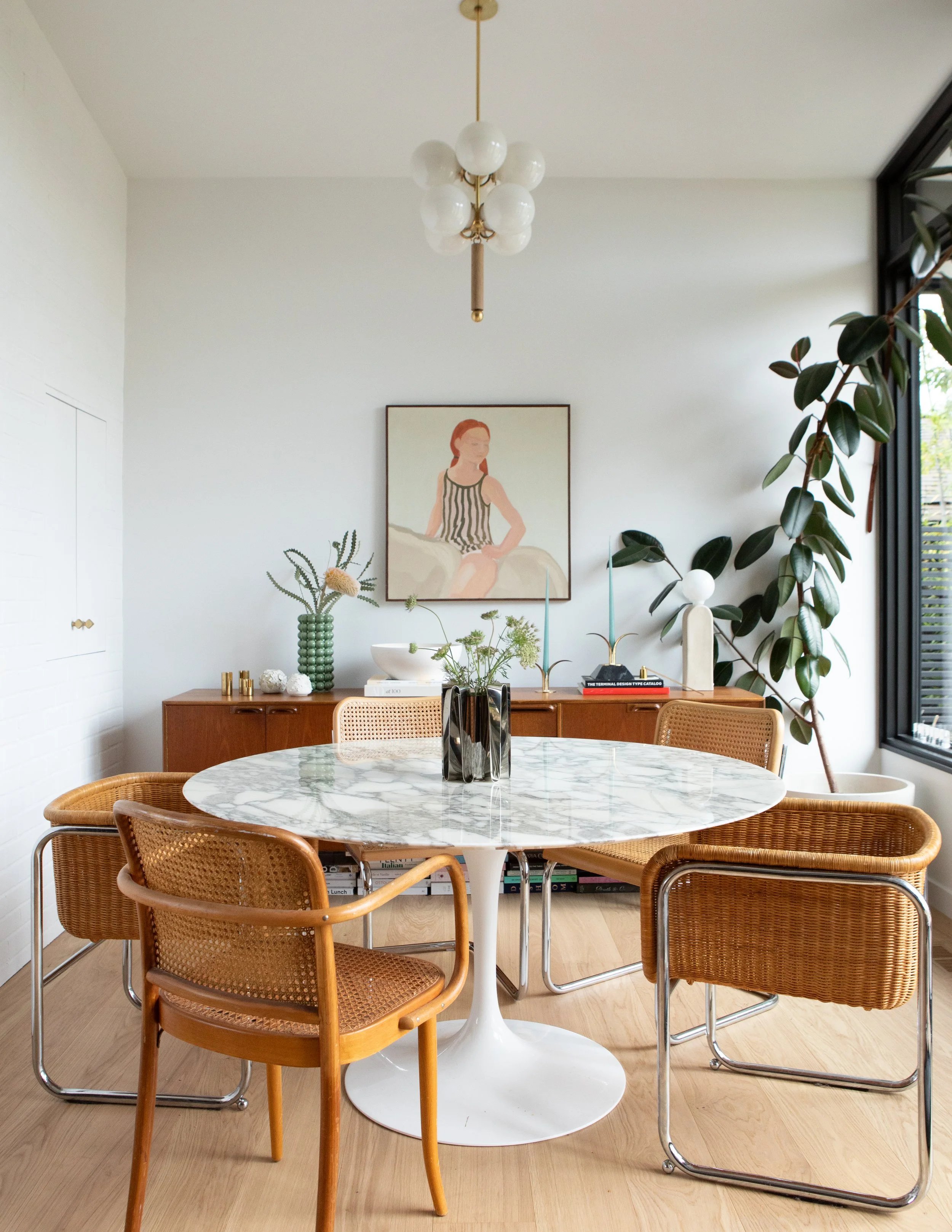 A dining room with a round white marble table, six rattan chairs, a wooden sideboard with vases and decor, with artwork on the wall, plants, and a window letting in natural light.