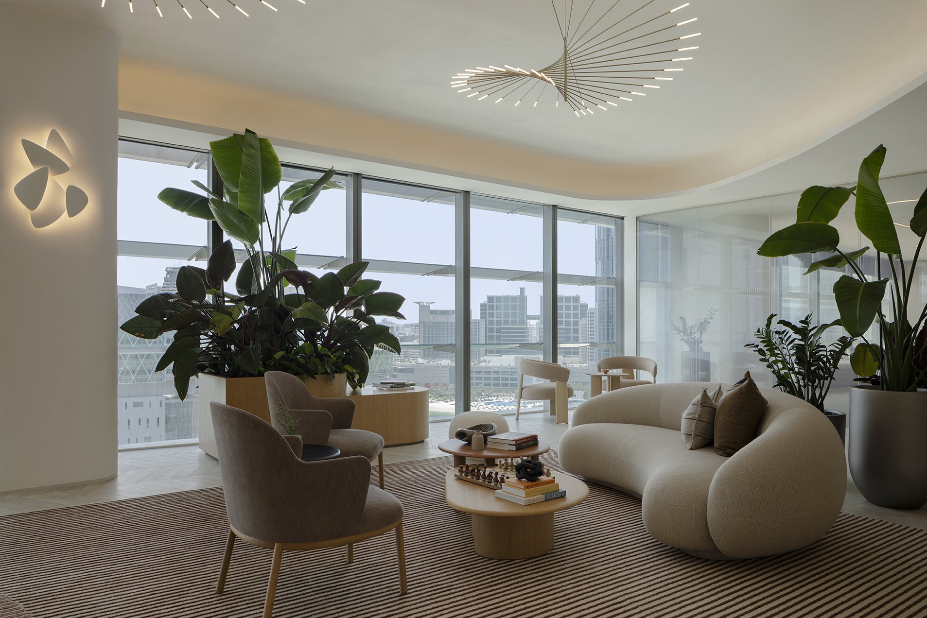 Modern living room with large windows, green plants, a beige sofa, and chairs, decorated with books and a chess game, in a city skyscraper.