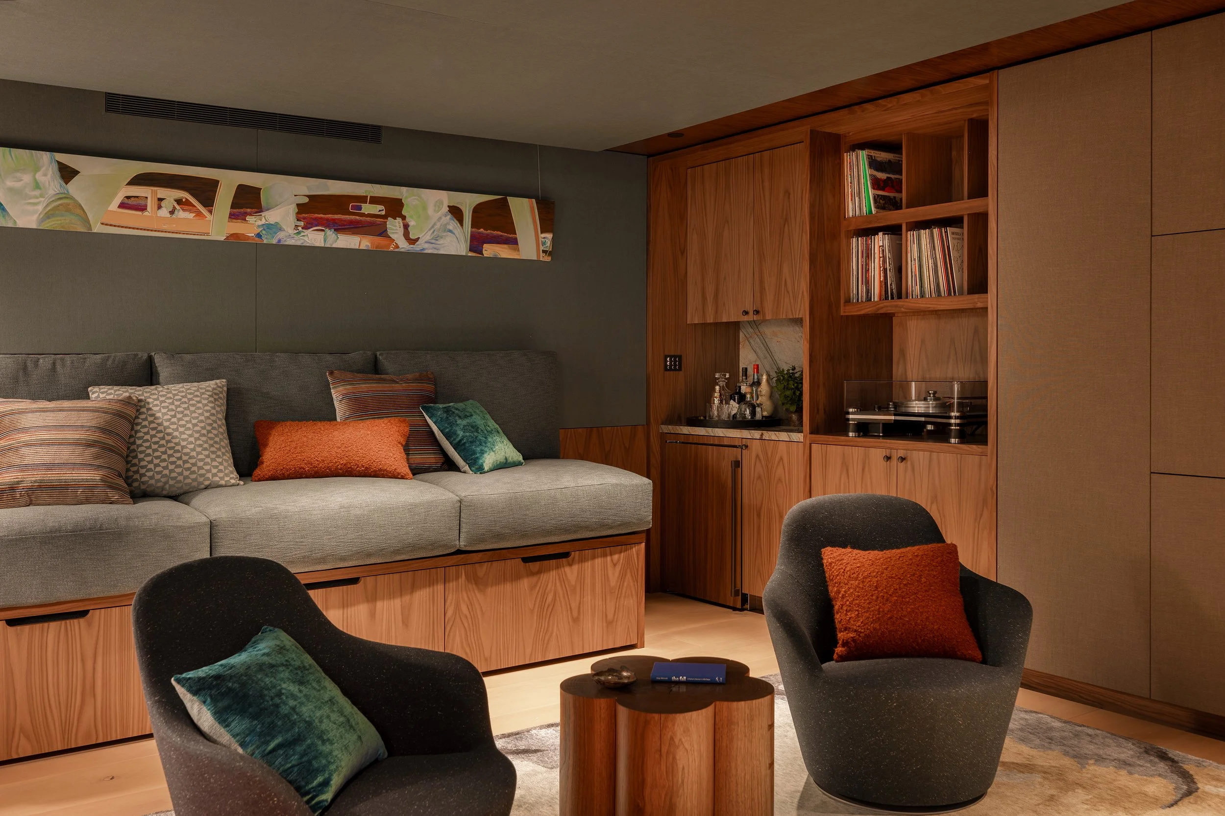 A cozy living room area with a gray sofa decorated with various colorful pillows, a black armchair with a green pillow, a wooden coffee table, and a built-in wooden bookshelf with vinyl records and beverages. Above the sofa is a long rectangular abst