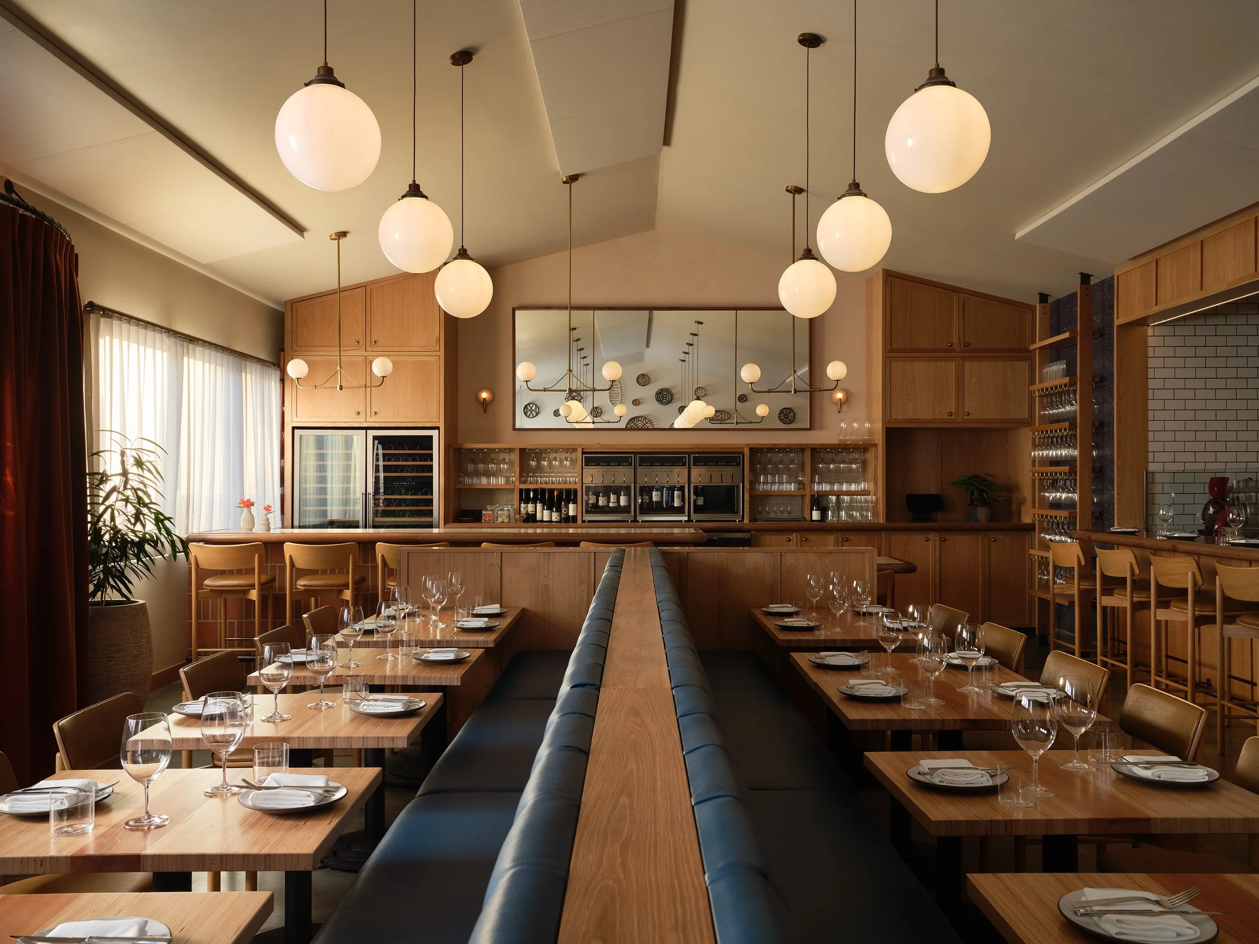 Interior of a restaurant with wooden tables set for dining, wine glasses, and white plates, pendant lights hanging from the ceiling, and a large mirror on the wall reflecting the decor and lighting.