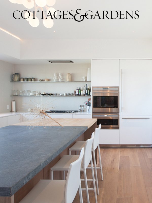 Modern kitchen with white cabinets, open shelves with dishes, built-in oven, and a kitchen island with four white chairs. A sparkler is on the island. The room has wood flooring and minimalist decor.