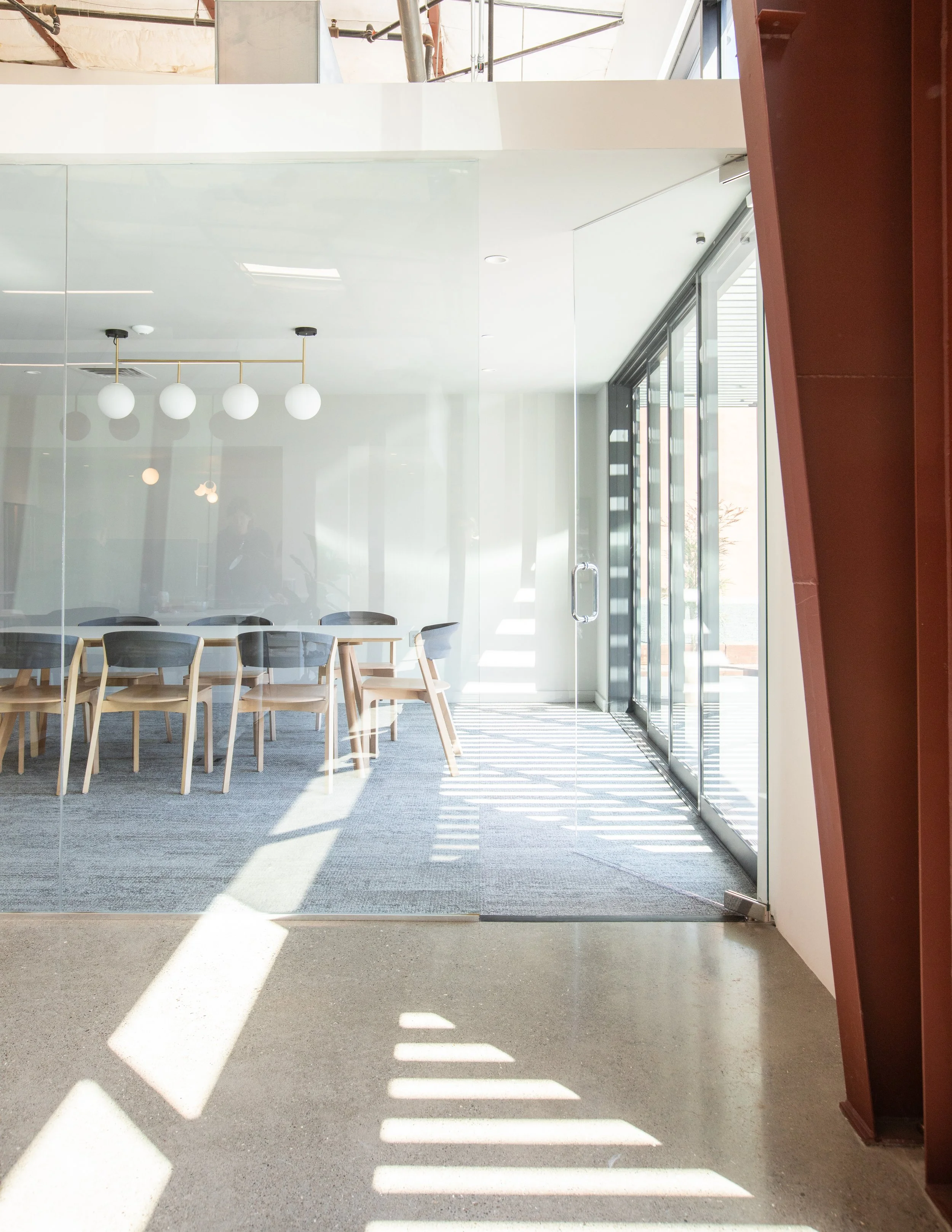 Interior view of a modern conference room with large glass windows, a white ceiling, a row of chairs, and hung spherical light fixtures, illuminated by natural sunlight.
