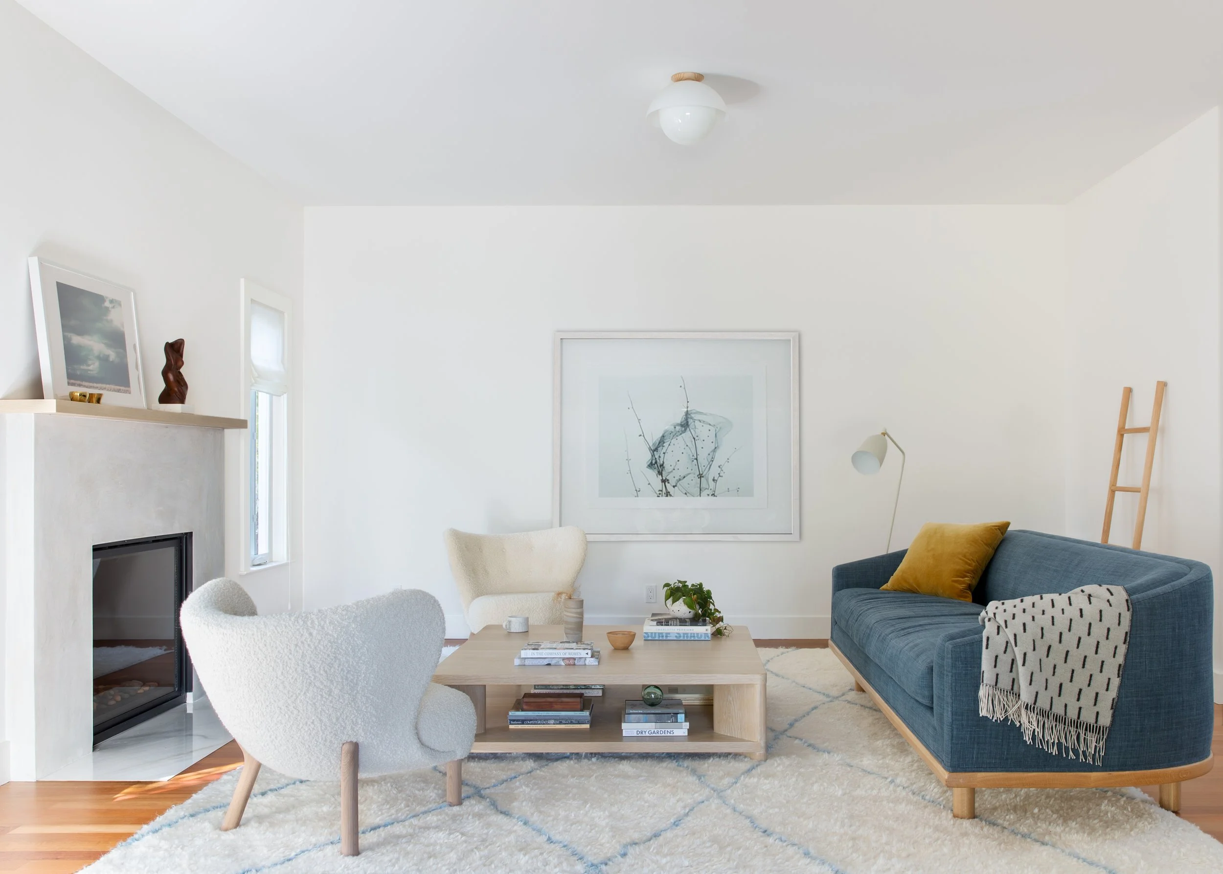 A cozy living room with white walls, a white ceiling, and wooden flooring. It features a blue sofa with yellow pillows, a white armchair, a wooden coffee table with books and decor, a framed abstract art piece on the wall, a white floor lamp, and a w