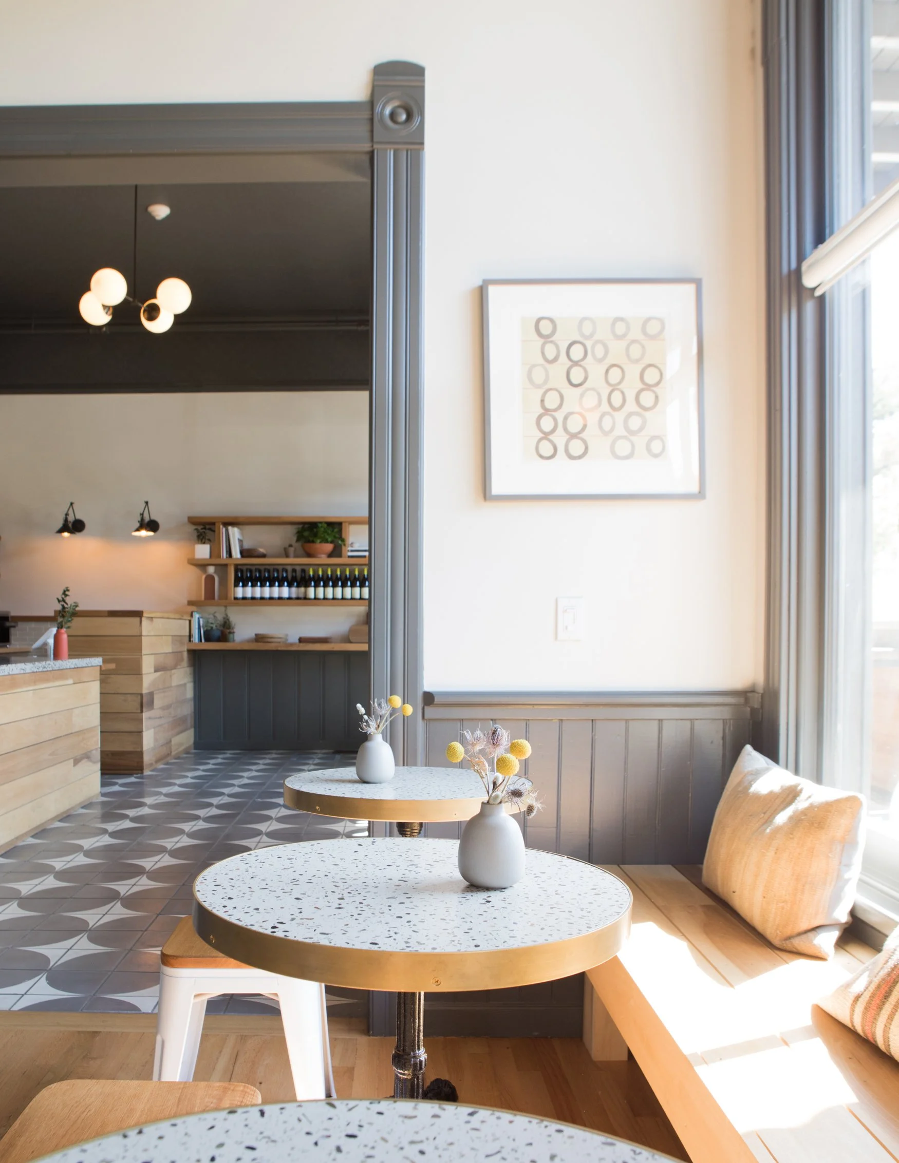 Interior of a modern café or dining area with white terrazzo tables, vases with dried flowers, a large window with curtains, and artwork on the wall. There is a bar skillfully stocked with wine bottles and decorated with plants.