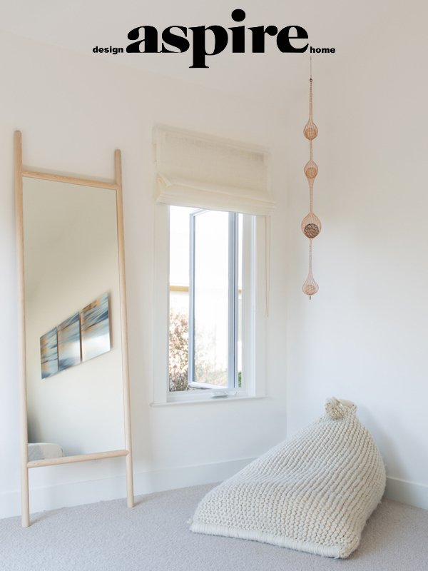 A minimalistic room with a large mirror leaning against the wall, a window with a white blind, a woven hanging decoration, and a knitted bean bag chair on a light-colored carpet.