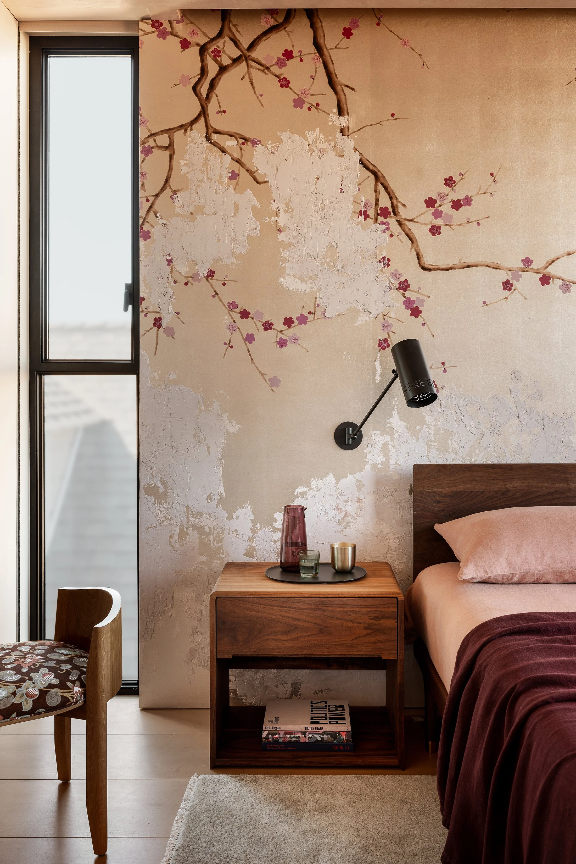 A bedroom with a floral and branch mural on the wall, a wooden nightstand with decorative vases and a newspaper, a bed with blush and dark burgundy bedding, next to a window, and a wall-mounted black reading lamp.