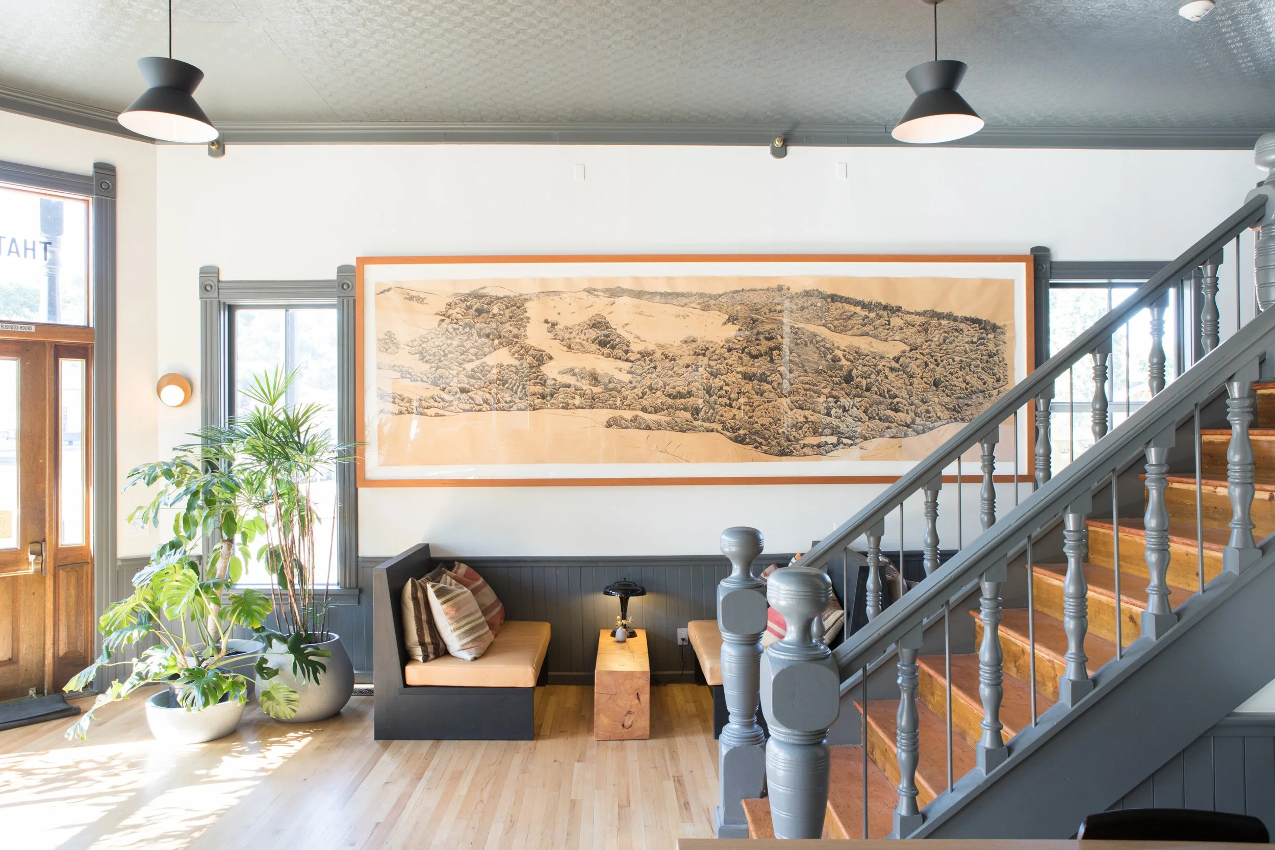 Interior of a cozy living room with light-colored hardwood floor, large windows, a framed landscape picture on the wall, a staircase with wooden steps and gray spindles, a plant in a large pot, and a small seating area with striped pillows.