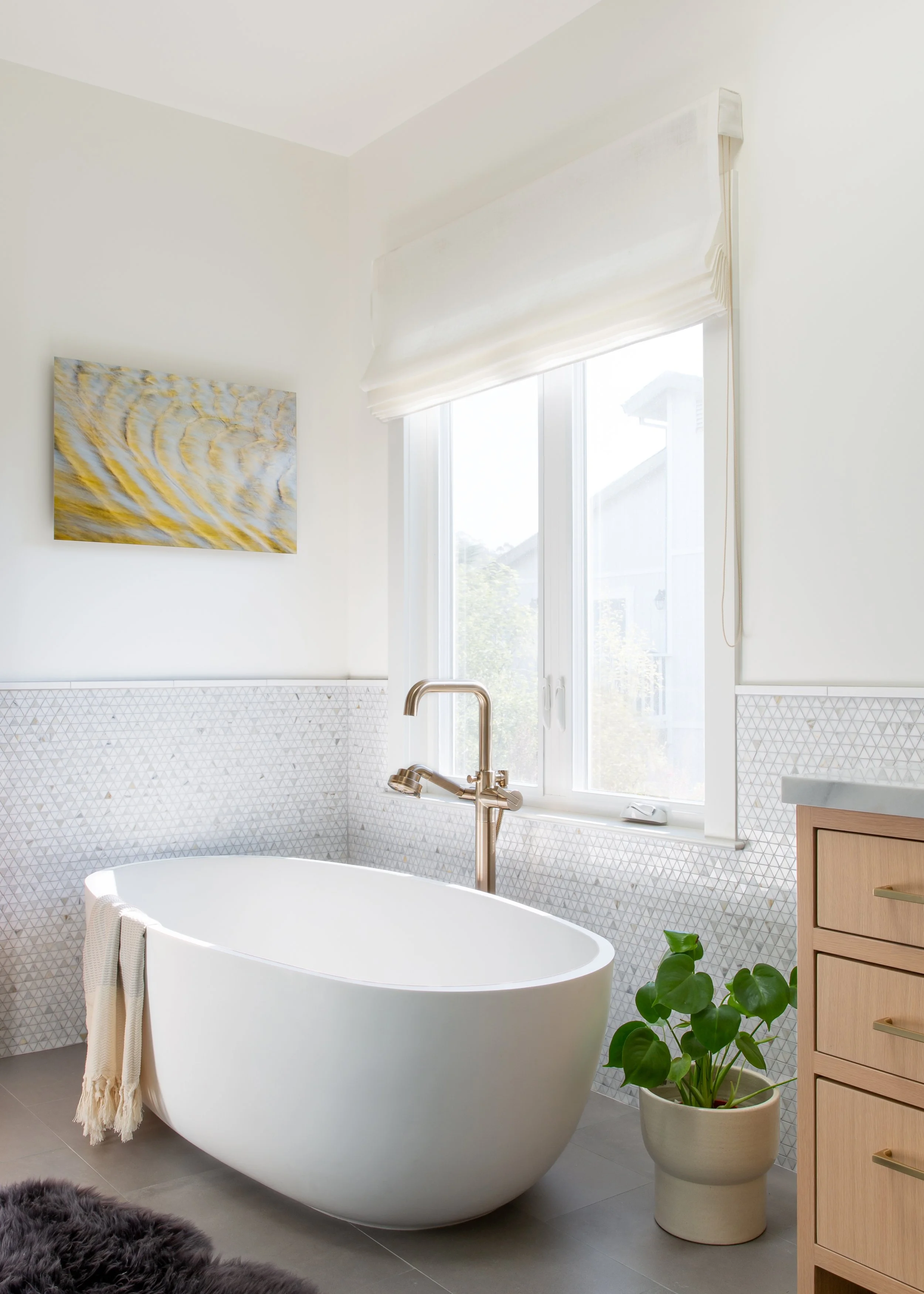 A modern, minimalist bathroom with a white free-standing bathtub, a gold-colored faucet, and a large window with white blinds. There is a colorful abstract painting on the wall, a potted green plant, a wooden cabinet with gold handles, and a gray rug