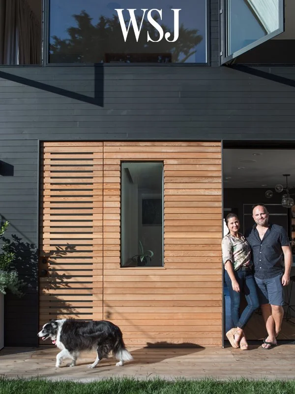 A modern house with black and natural wood exterior, featuring a door with a wooden slat design and a narrow window. Two people stand at the doorway, smiling, with a dog walking on the grass outside. The WSJ logo is at the top of the image.