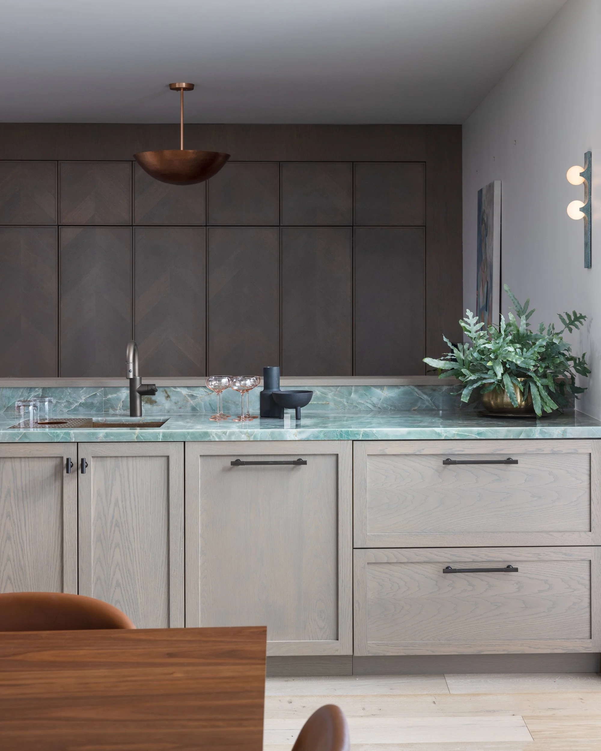 Modern kitchen with beige wood cabinets, green marble countertop, black faucet, and a large potted plant, with gray upper cabinets and wood flooring.