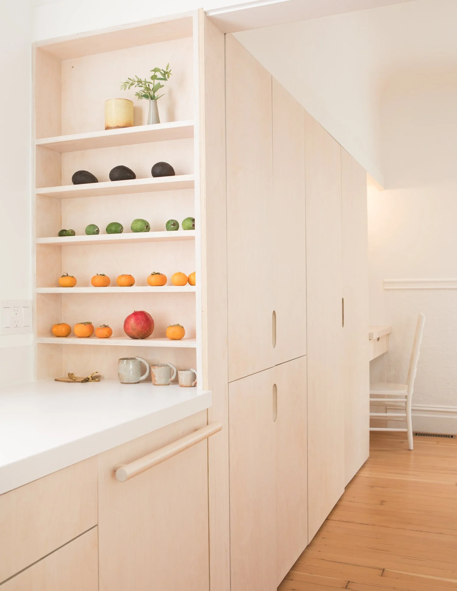 A kitchen with open shelves displaying fruits, potted plants, and kitchenware, with a white cart and a chair nearby, all with light wood finishes and a hardwood floor.