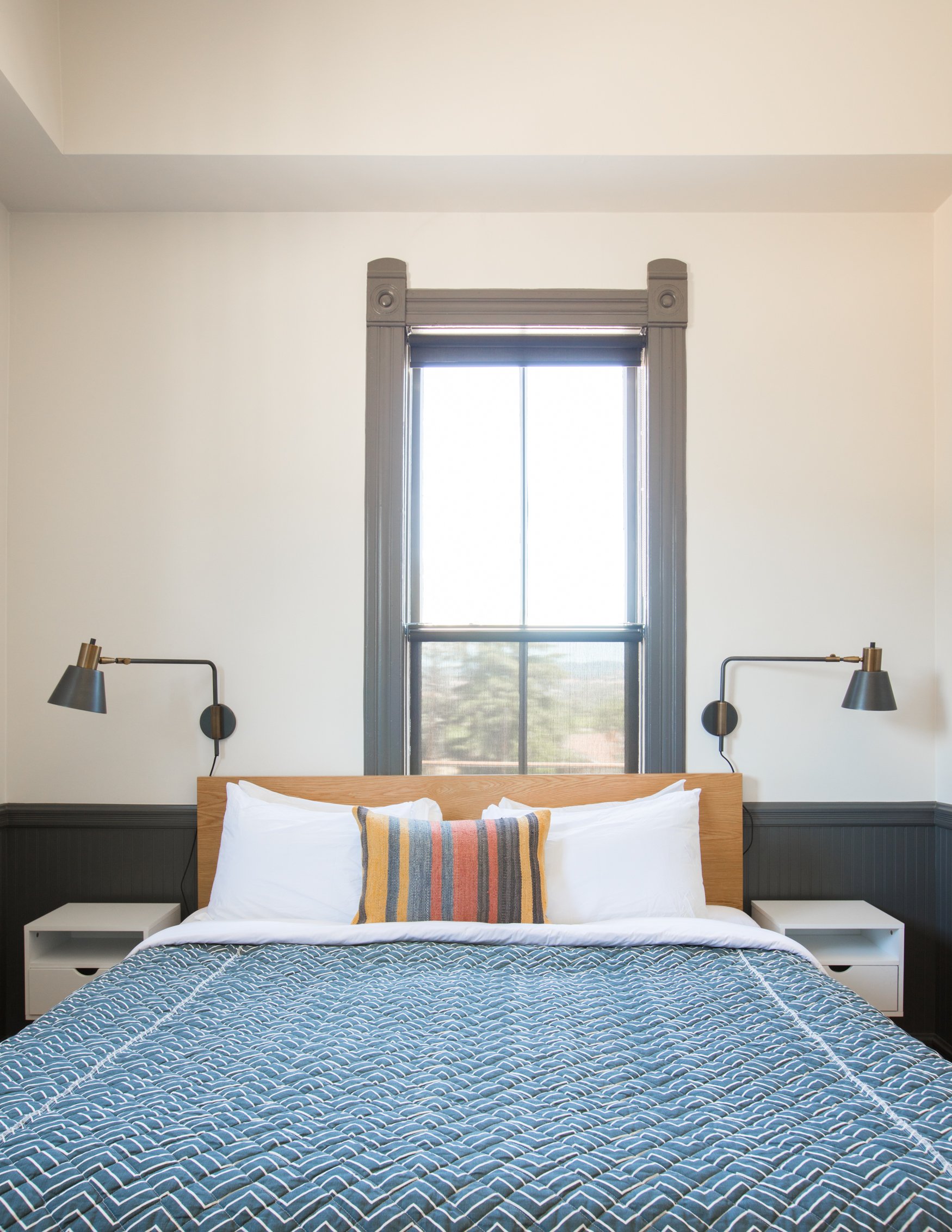 A neatly made bed with white pillows, a colorful striped pillow, and a patterned blue bedspread in front of a large window with gray trim and a black shade, flanked by two black wall-mounted reading lamps.
