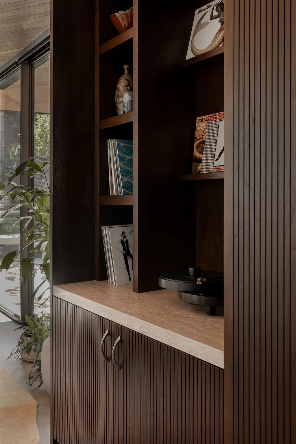 Close-up of a bookshelf with magazines and decorative items, partially obscured by a wooden door with vertical slats, in a room with large glass windows and a plant to the side.