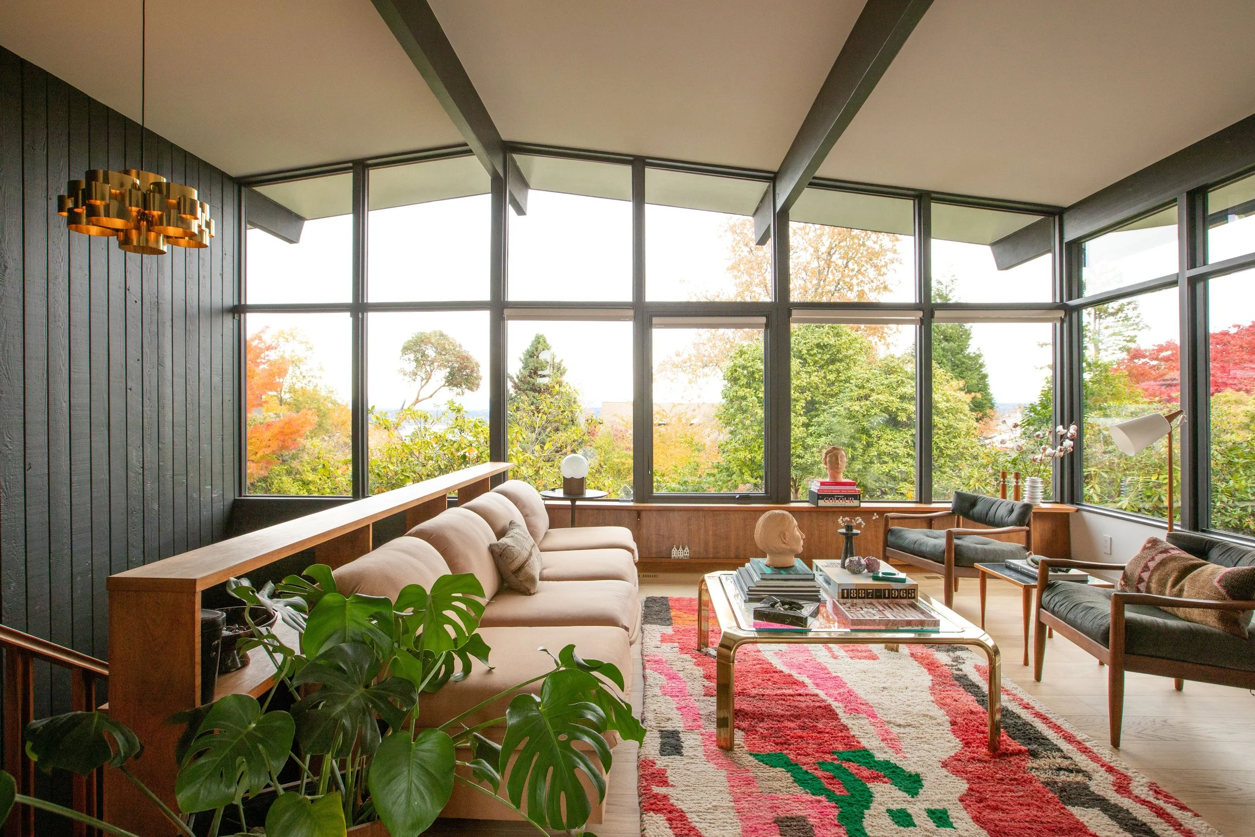 Living room with large glass windows showing a view of trees with autumn foliage, modern furniture including a beige sofa, black and wooden chairs, colorful geometric rug, and decorative items on a coffee table.