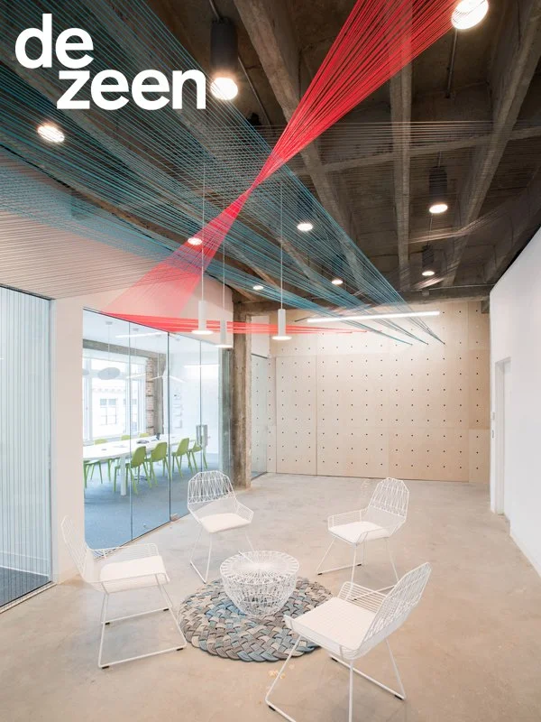 Modern office lounge with white wire chairs around a woven rug, glass walls, and decorative red and blue string art fixtures hanging from the ceiling.