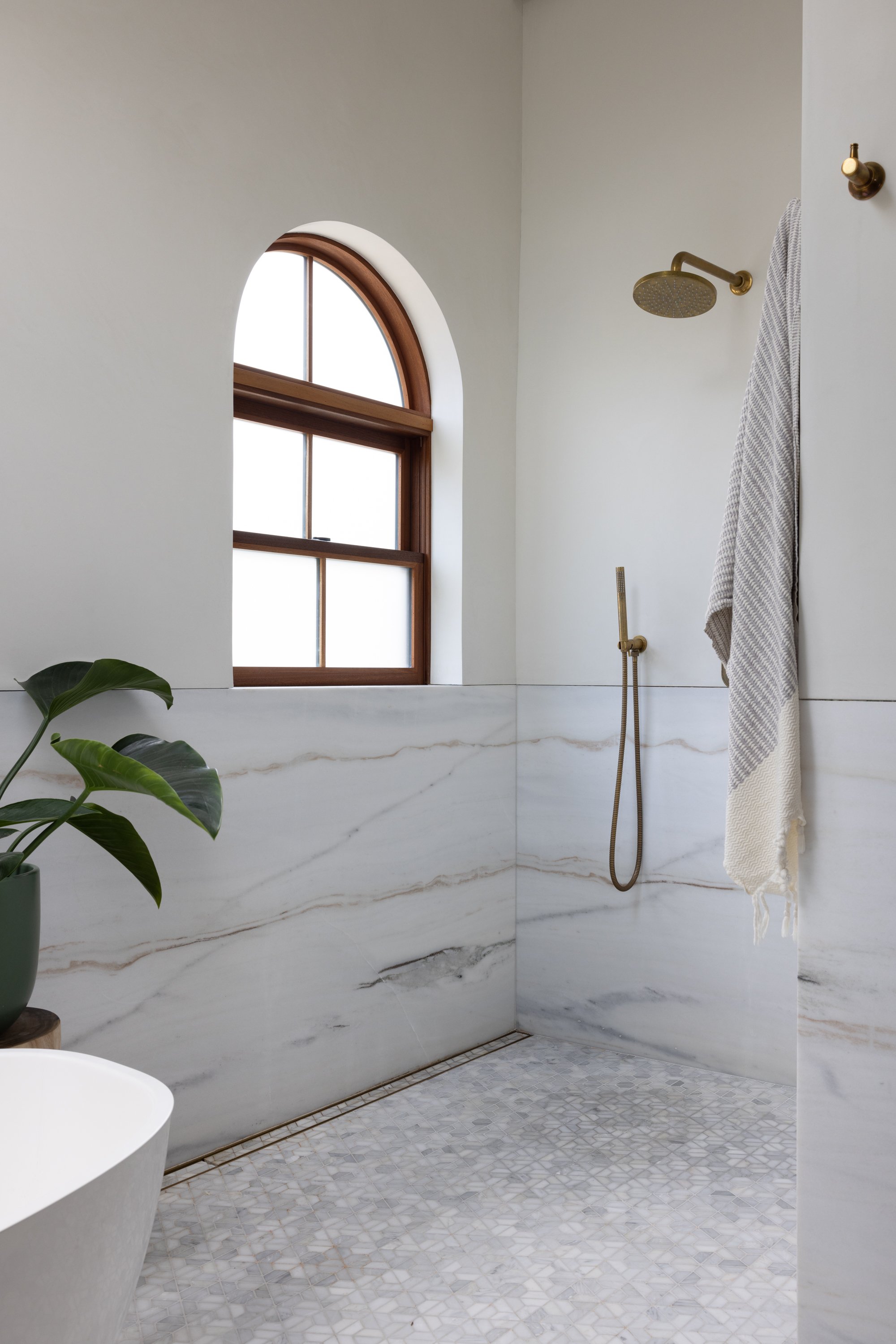 A modern bathroom with a large arched window, a marble wall with veining, a rainfall showerhead, a handheld shower, a hanging towel, and a green potted plant.