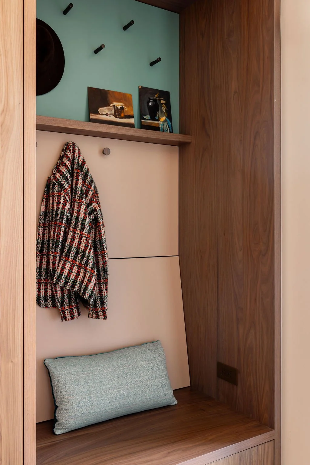 Wooden entryway nook with coat hooks, a colorful patterned coat hanging, a cushion on the floor, and small paintings on a top shelf.