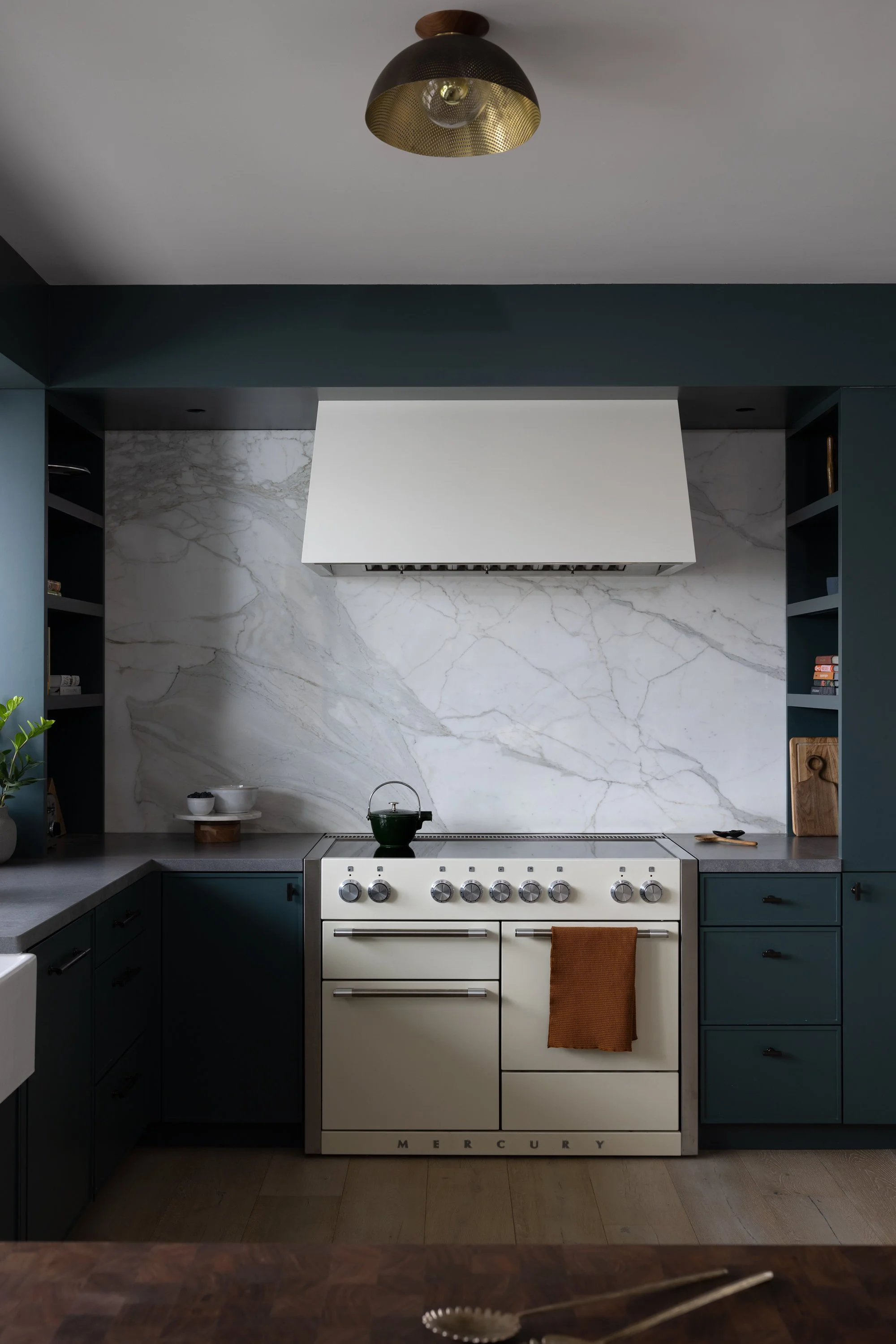A modern kitchen with a white vintage Mercury stove, dark green cabinets, a marble backsplash, and open shelving with small decorative objects, seen from a dining area with wooden flooring and utensils in the foreground, and a ceiling light fixture.