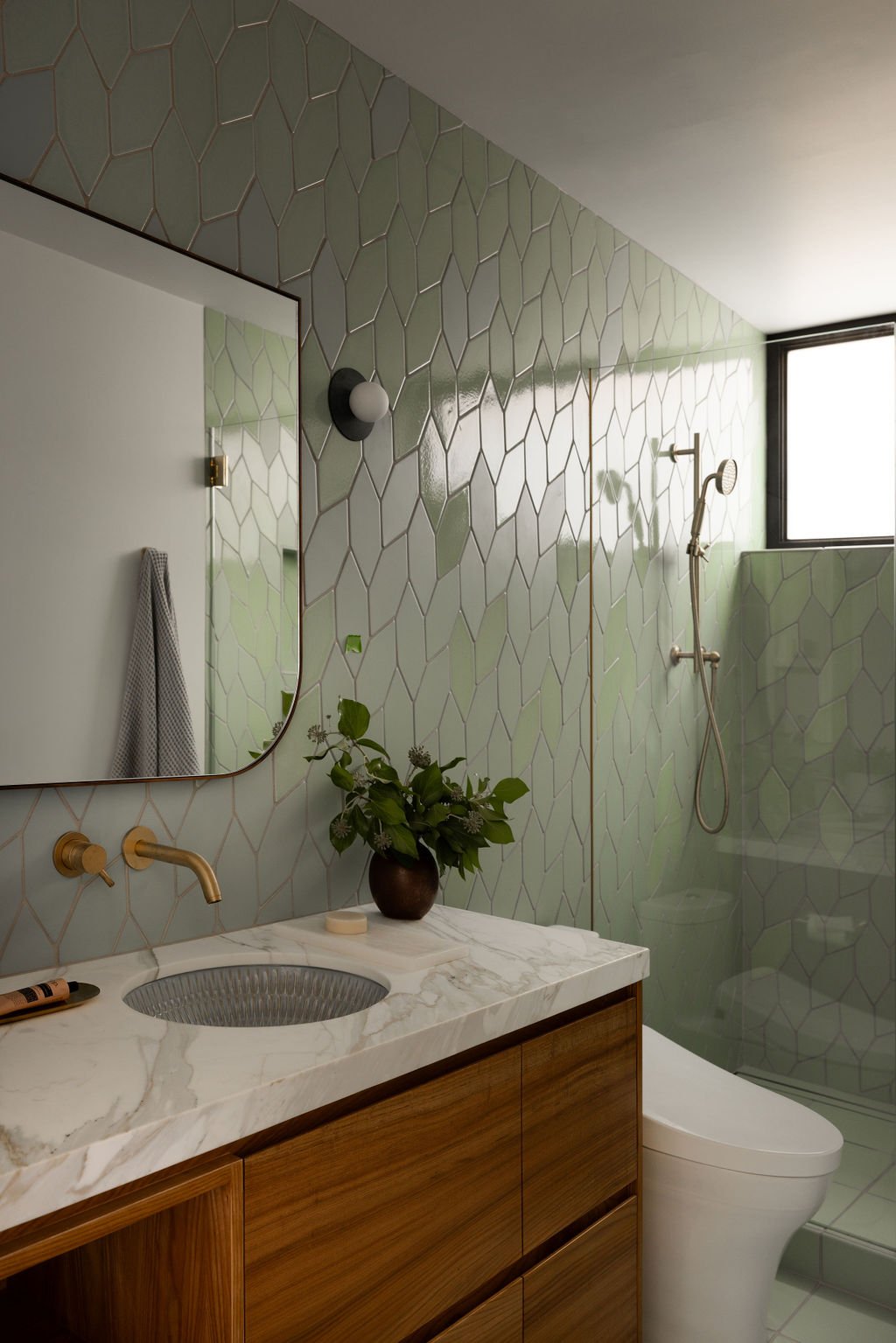 Modern bathroom with green tiled walls, a marble countertop sink, wooden vanity, a mirror, a plant in a brown pot, a wall-mounted light fixture, and a walk-in shower with a window.