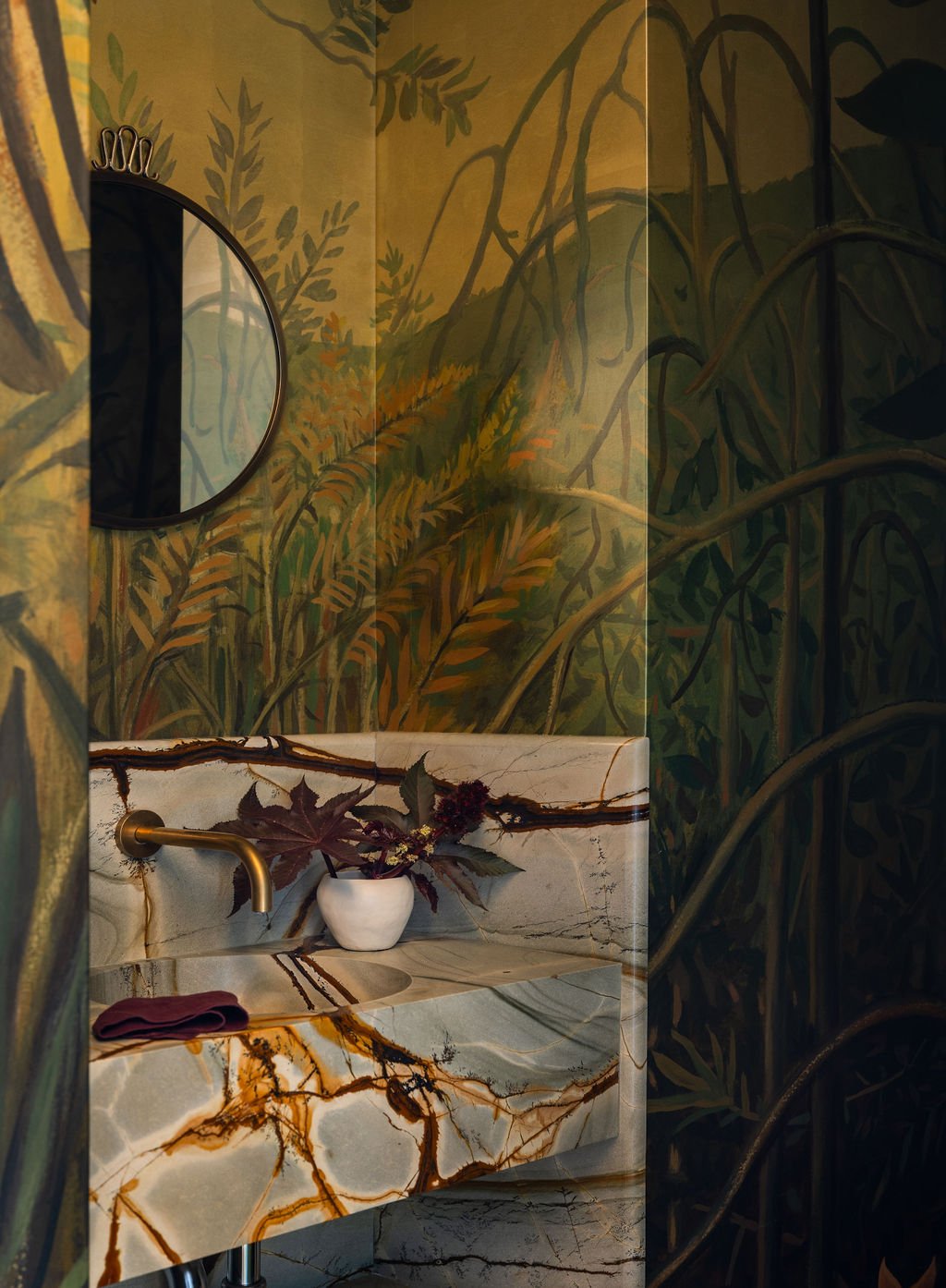 Bathroom with nature-themed mural depicting forest and foliage, marble sink with earthy vein patterns, a small white vase with dark leaves, and a round mirror on the wall.