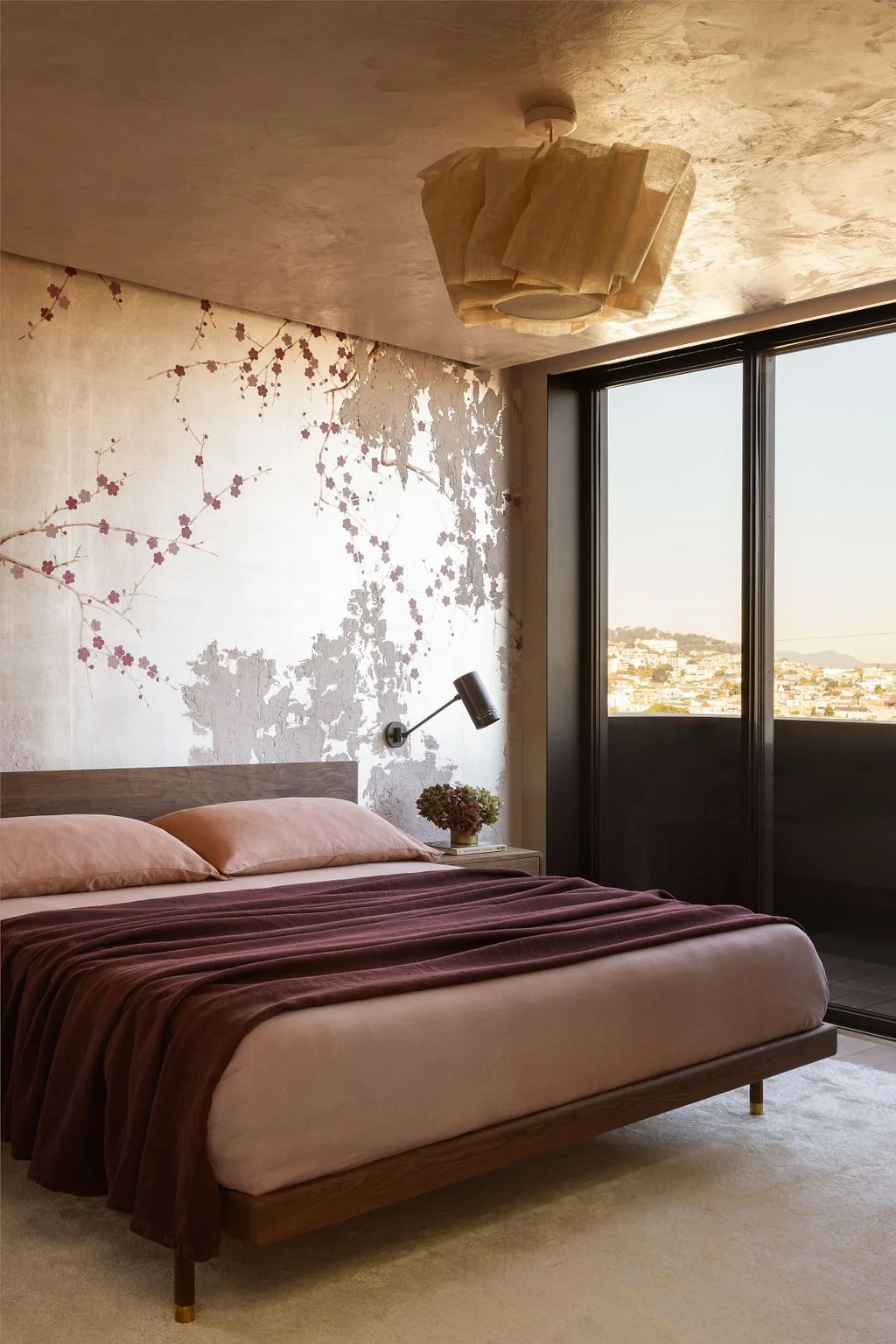 A bedroom with a wooden bed, pink pillows, and a purple blanket. A wall with a cherry blossom mural, a black wall-mounted lamp, and a small table with flowers. Window with a view of a city and distant hills.