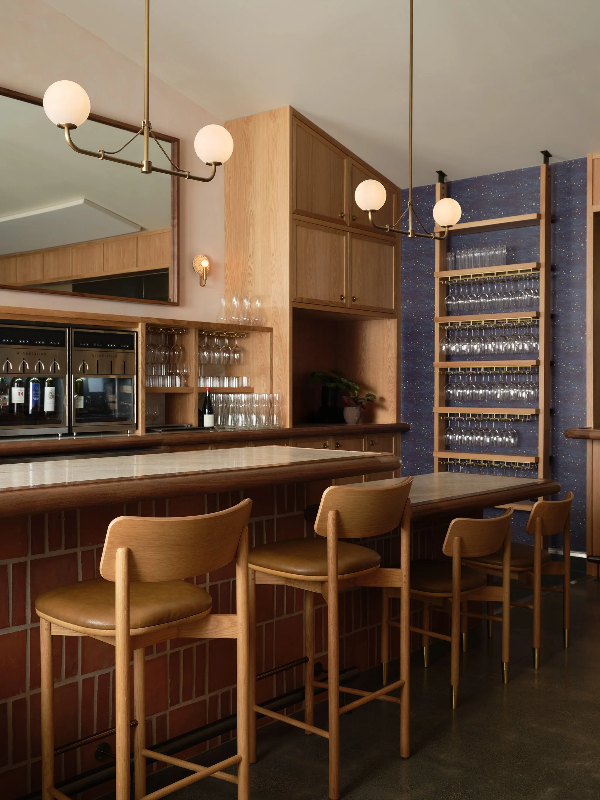 A bar area with a counter, wooden bar stools, a mirror, wall lights, wooden cabinets, and a wine glass rack against a dark starry sky painted wall.