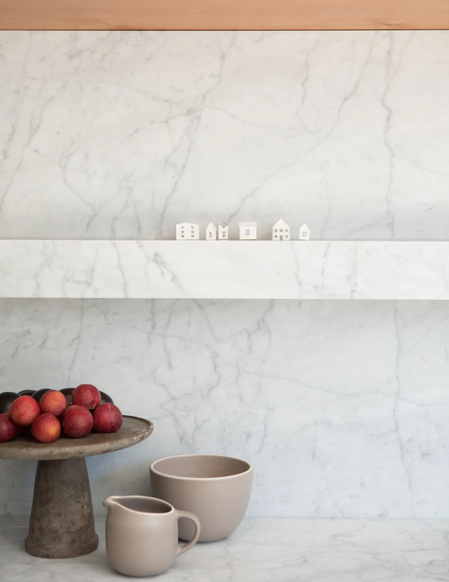 A marble countertop with a small white ceramic pitcher, a white bowl, a concrete pedestal holding a cluster of red apples, and a marble shelf with small white house-shaped decorations.