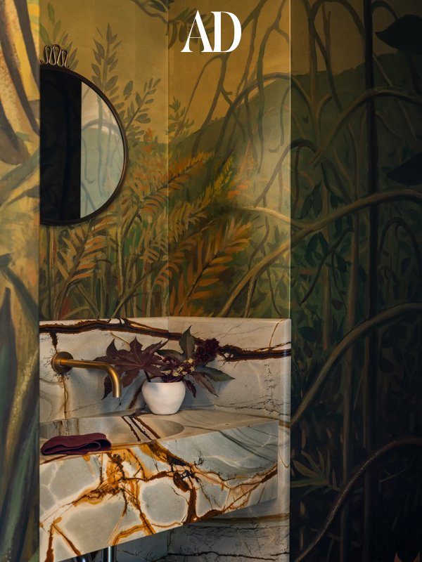 A luxurious bathroom with a forest scene painted on the walls, a marble sink with gold accents, and a small white vase with purple plants on the countertop.