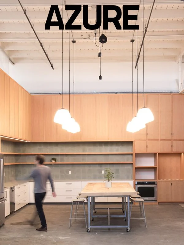 A modern kitchen with wooden cabinets, open shelving, and a central island with chairs. Lighting fixtures hang from the ceiling, and a person is walking past the island.