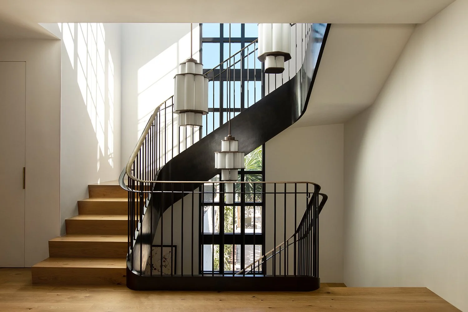 Modern staircase with wooden steps, black metal railing, and hanging pendant lights in a bright, minimalistic interior with large windows and light-colored walls.