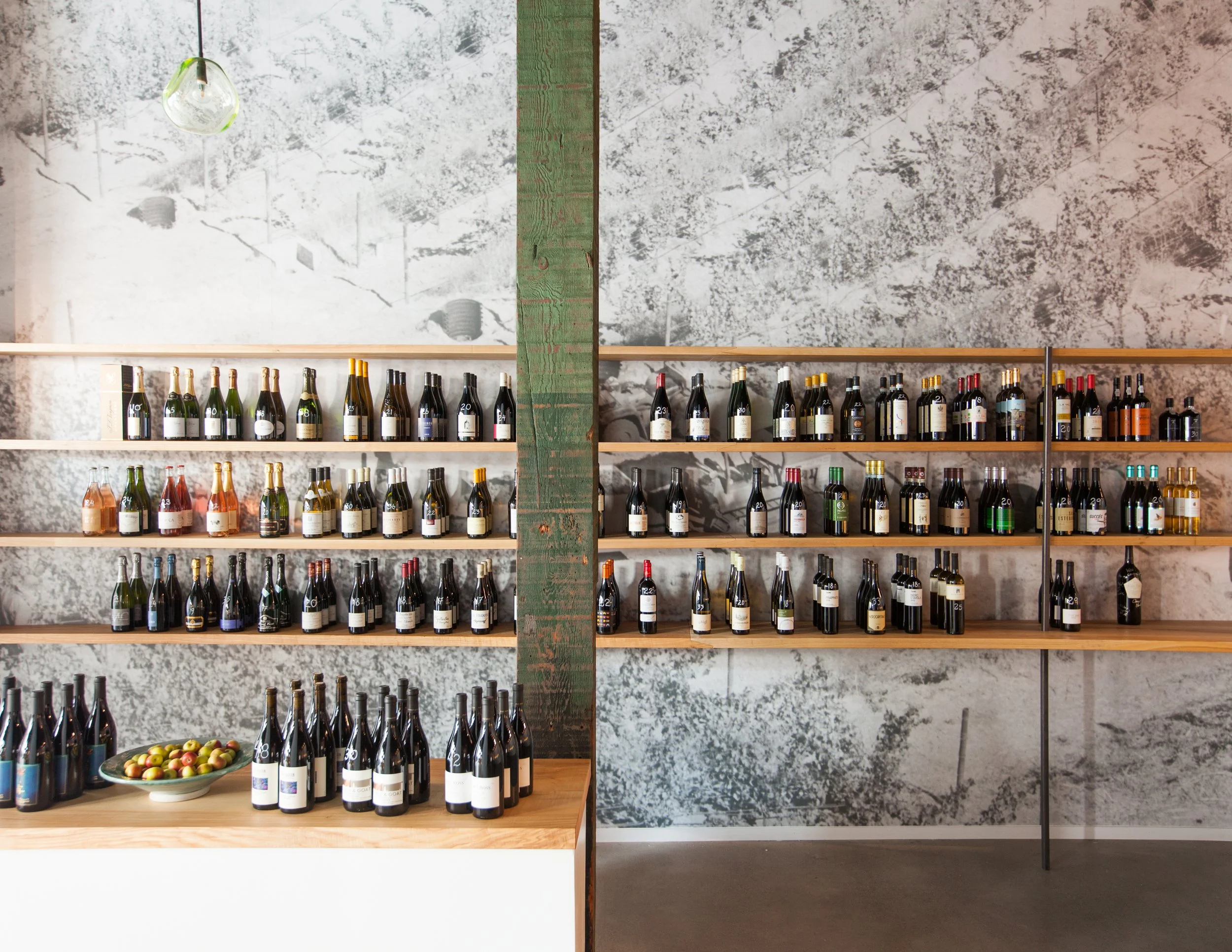 Interior of a store with wooden shelves filled with bottles of wine, decorated with a large gray and white textured wall and a green wooden post in the center.
