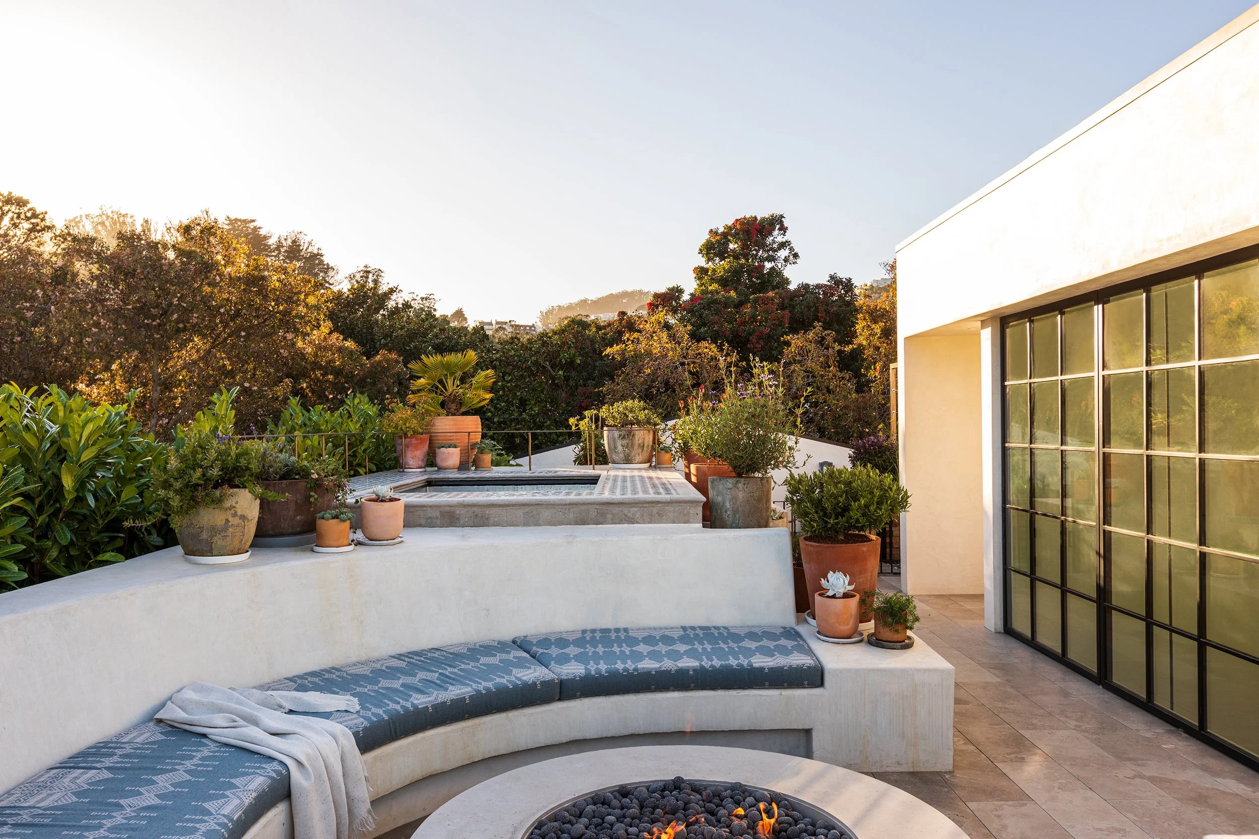 A modern outdoor terrace featuring a built-in curved seating area with blue patterned cushions, potted plants, and a fire pit with burning flames, surrounded by lush greenery and trees under a clear sky.