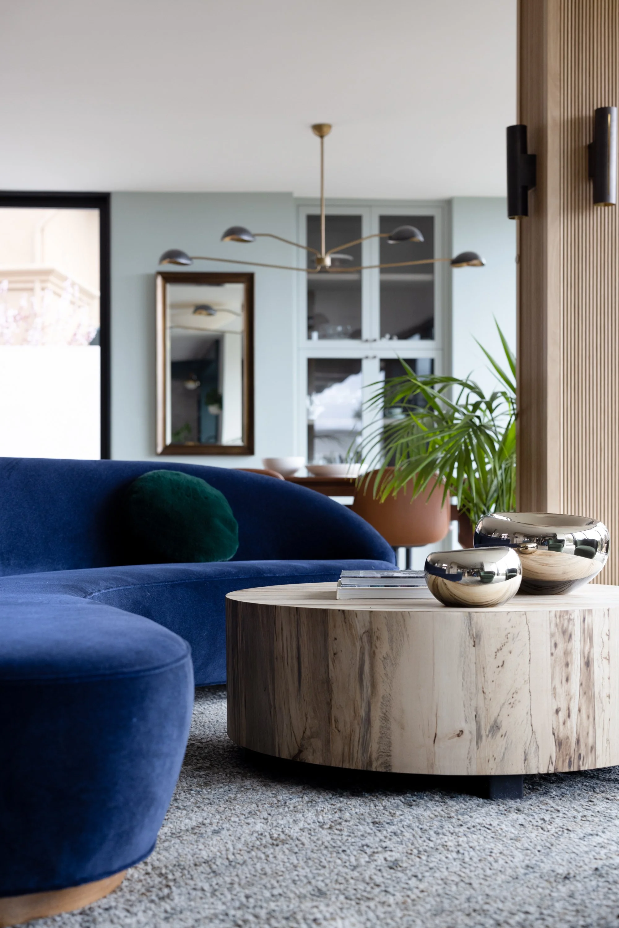 Living room with a blue velvet sofa, wooden coffee table with metallic bowls, green cushion, potted plant, mirror, wall-mounted speakers, and modern lighting fixtures.