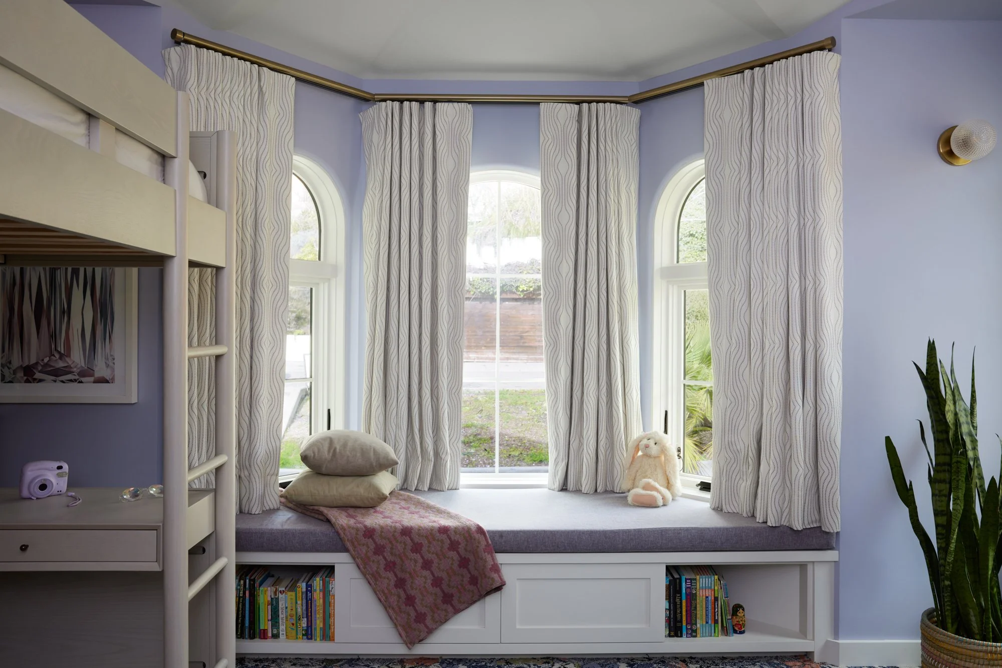 A cozy window nook with three large arched windows with patterned curtains, a small bed with pillows, a stuffed bunny, a pink blanket, bookshelves below, and a plant on the right.