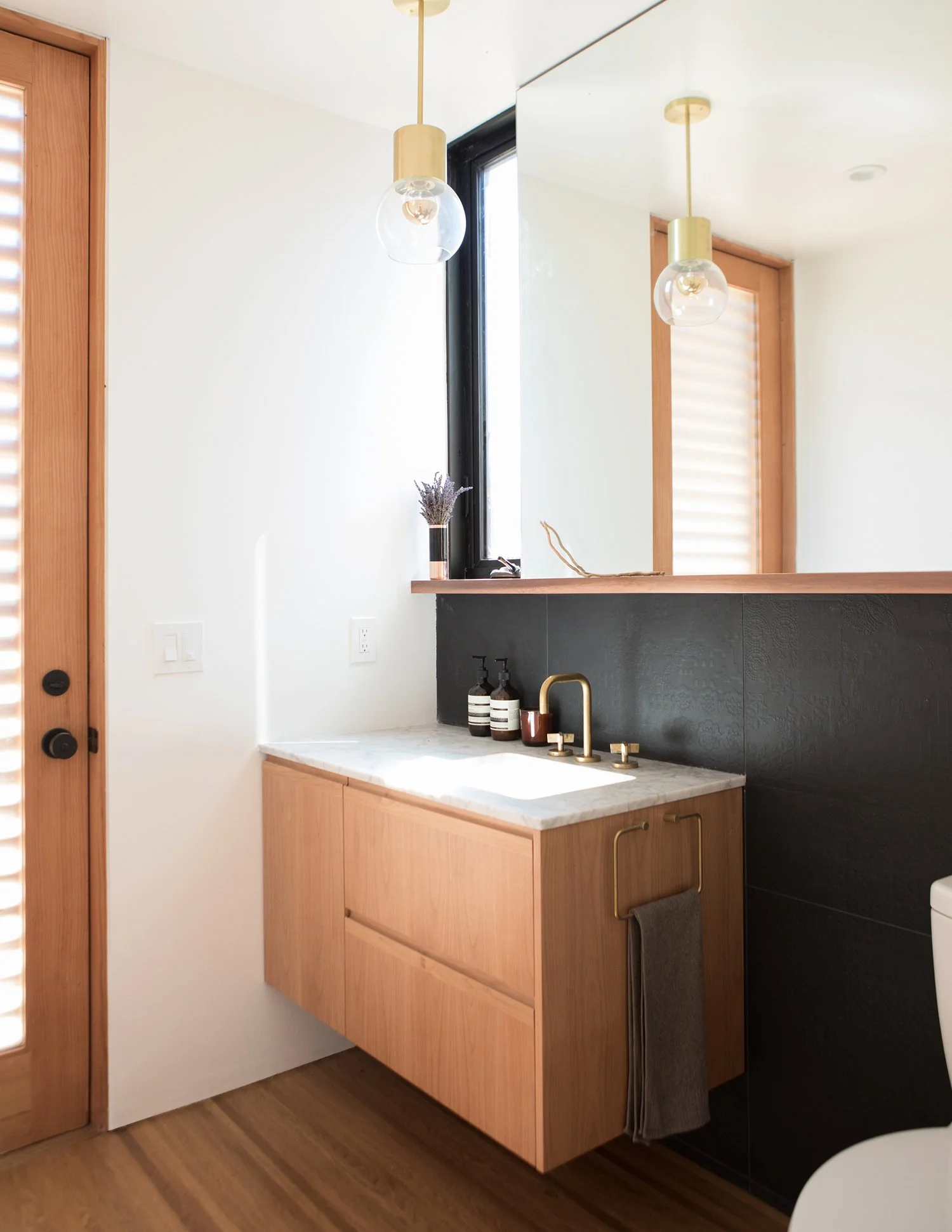A modern bathroom vanity with a black textured backsplash, wooden cabinets, a marble countertop, and a gold faucet. There is a small window, a mirror, and a light fixture hanging from the ceiling.