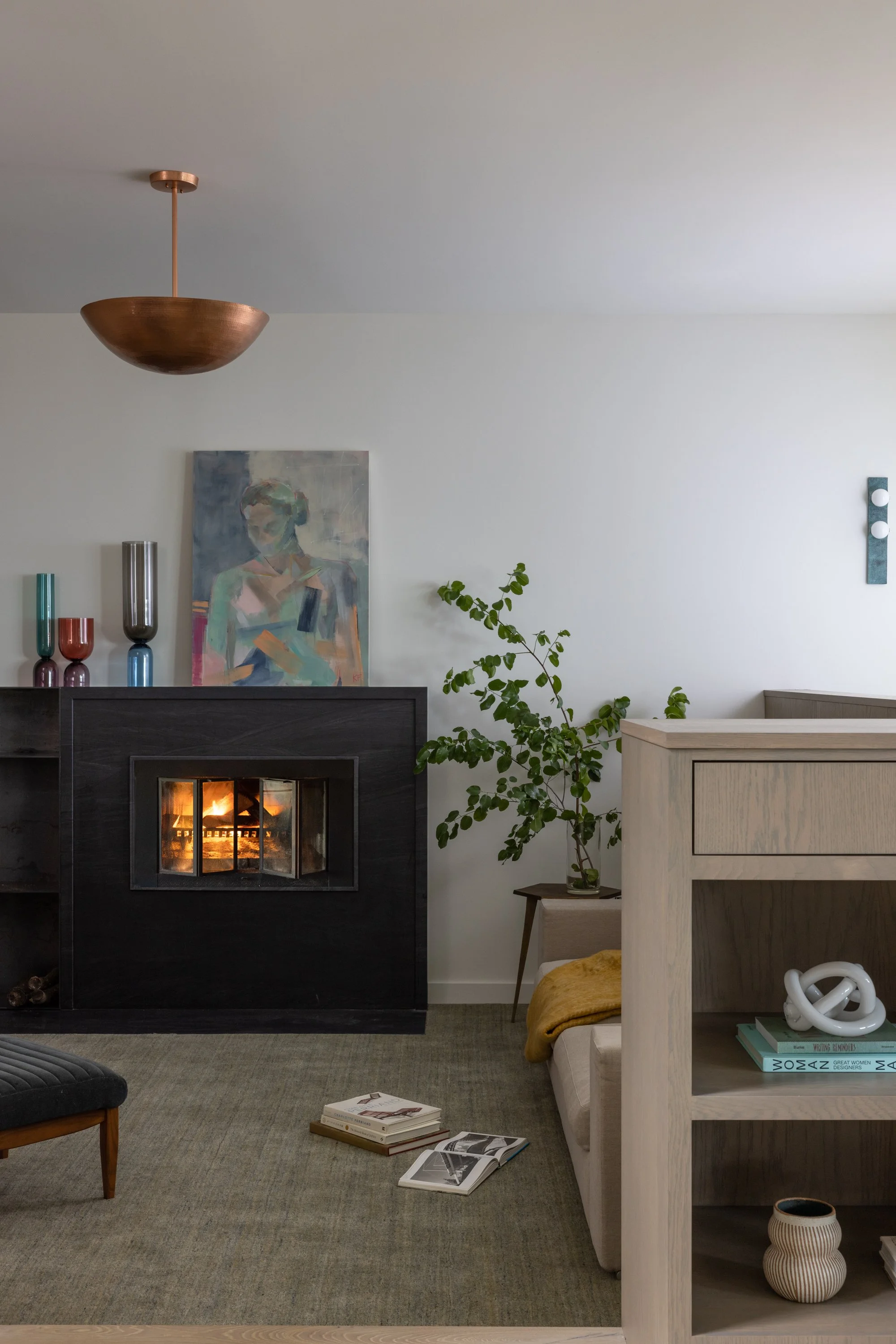 Living room with a modern fireplace, abstract portrait painting, colorful vases, green plants, a black ottoman, a beige sofa with a yellow blanket, open magazines, and a light-colored wooden shelving unit with decorative objects and books.