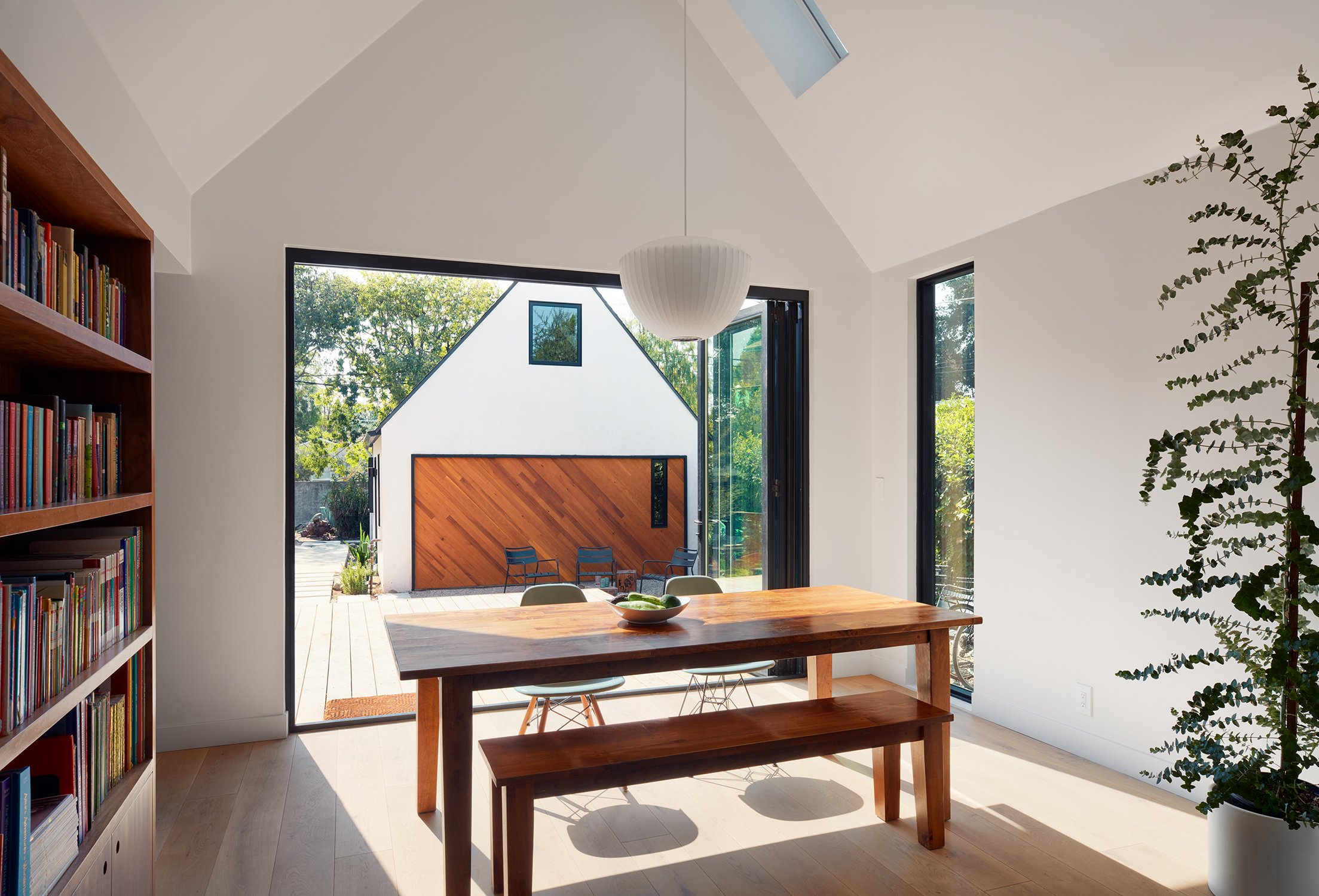 Interior view of a modern home with large windows, a wooden dining table with chairs, a ceiling light fixture, a bookshelf filled with colorful books, and a sliding glass door leading to an outdoor patio with seating and greenery.
