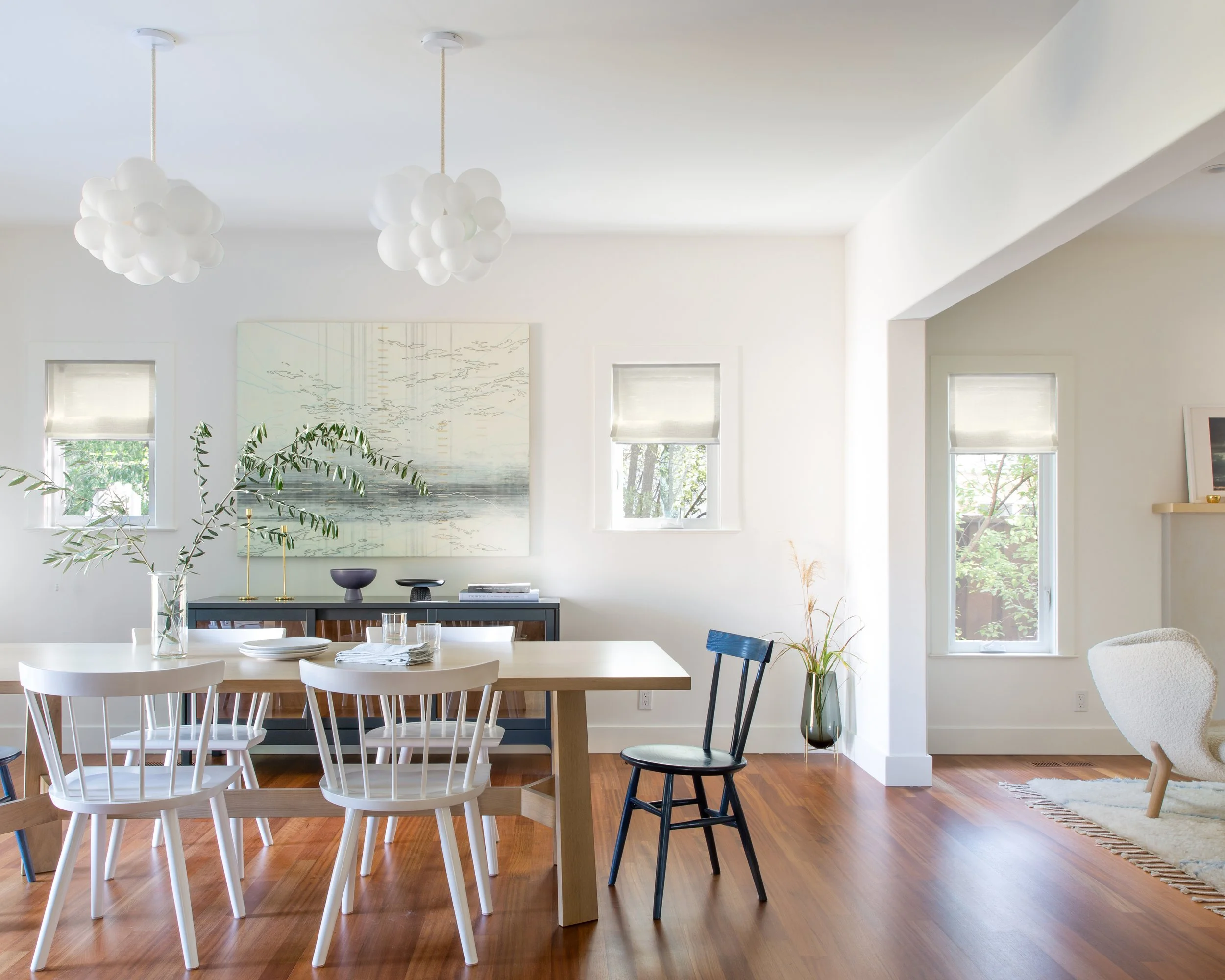 Bright, modern dining area with a wooden table surrounded by white chairs, a black chair, and contemporary decor including framed artwork, potted plants, and unique suspended light fixtures.