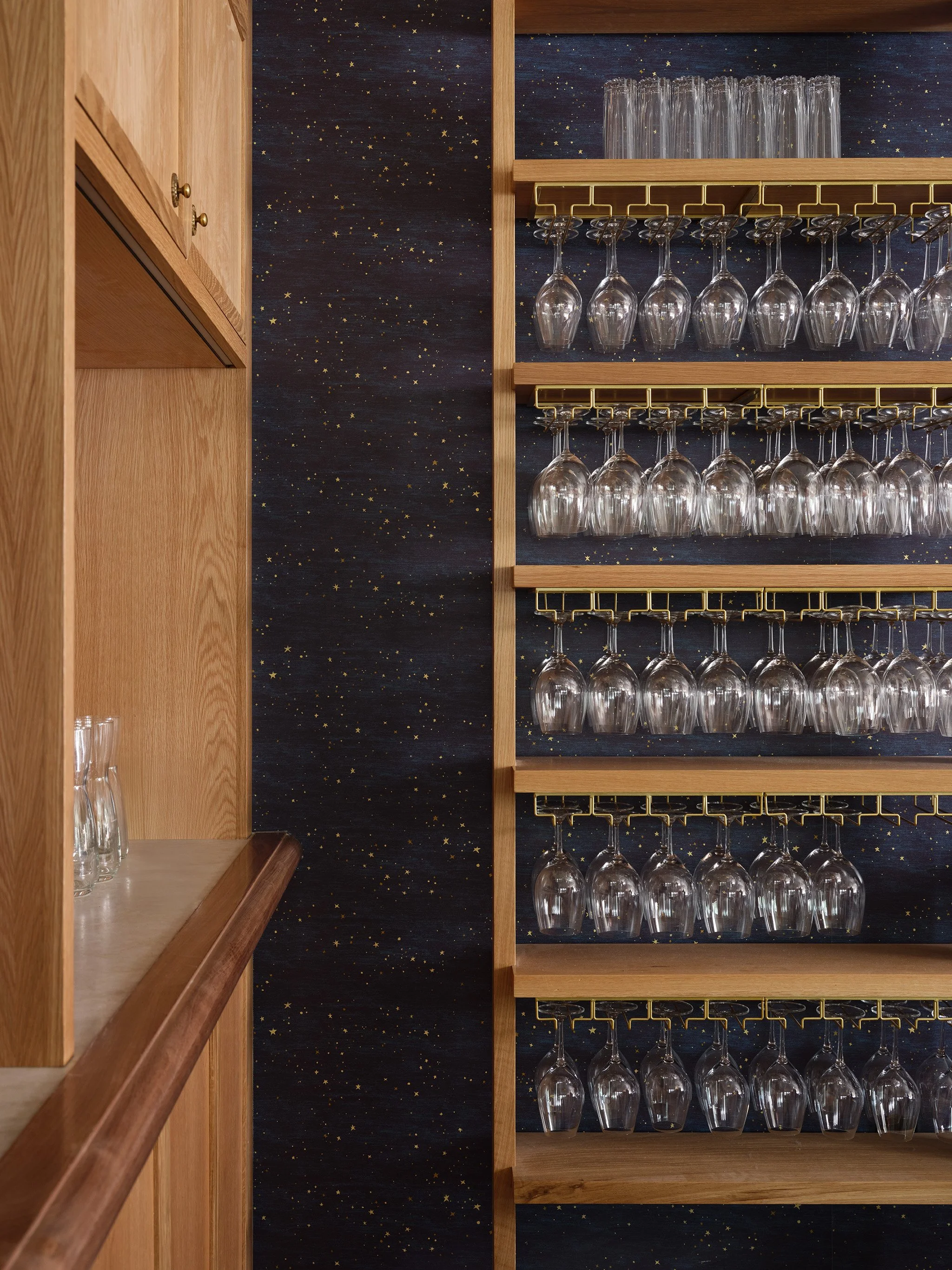 A wooden shelf with glasses hanging upside down on gold racks, against a dark blue wall decorated with small gold stars.