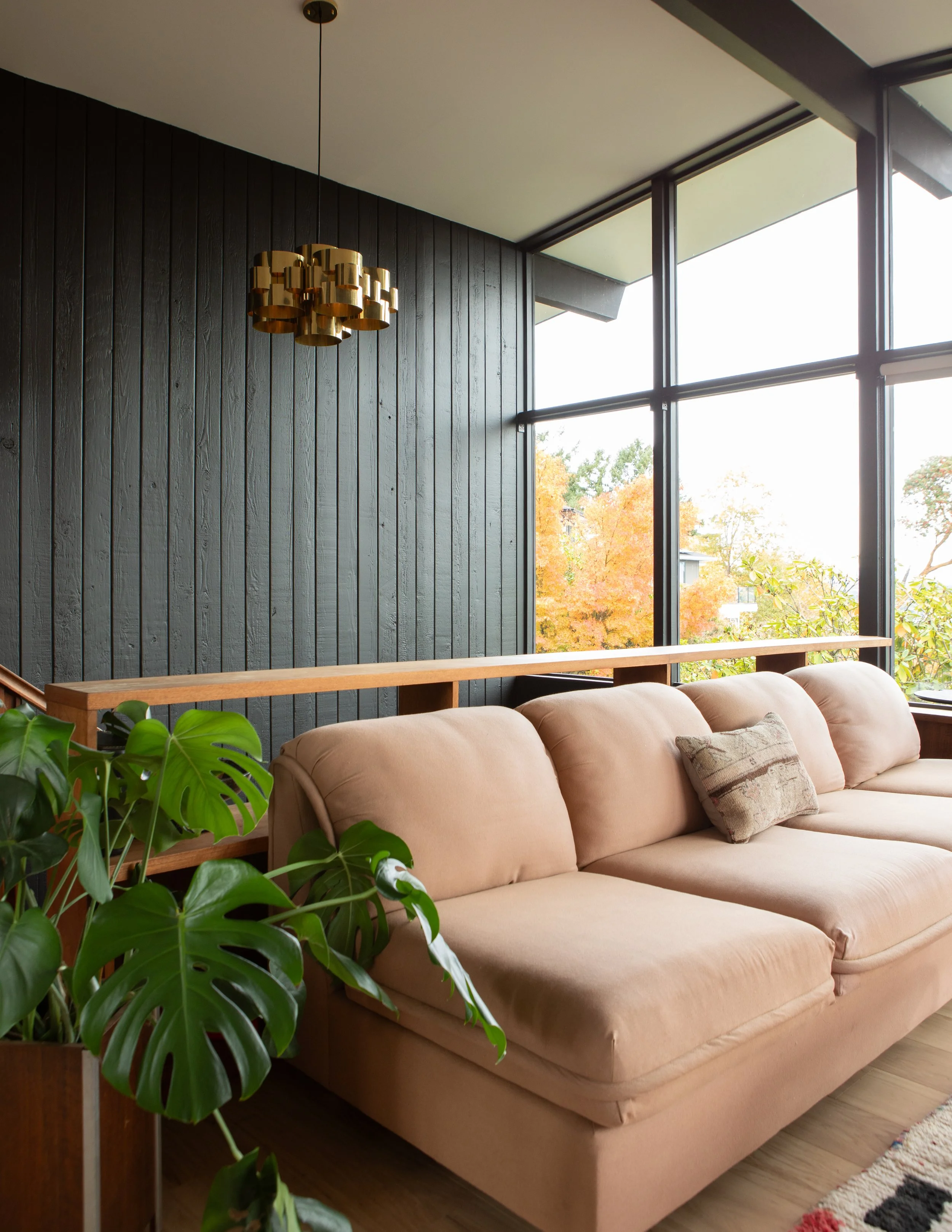 Living room with beige sofa, large windows showing autumn trees outside, dark wood-paneled wall, modern gold ceiling light fixture, green houseplant, and a colorful rug.