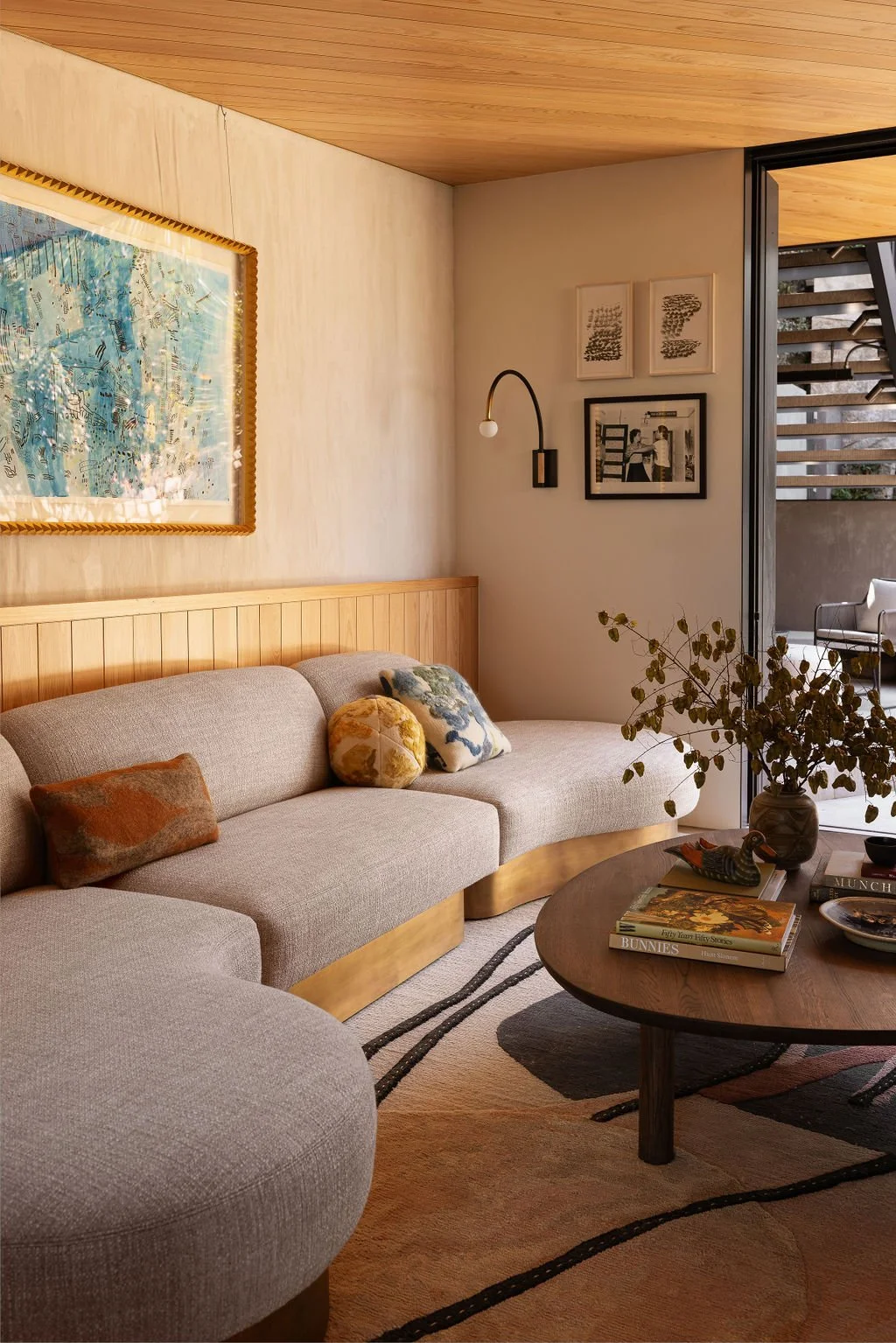 Living room with beige sofa, wooden coffee table, decorative pillows, framed artwork, wall-mounted light fixture, and open doorway to an outdoor patio.