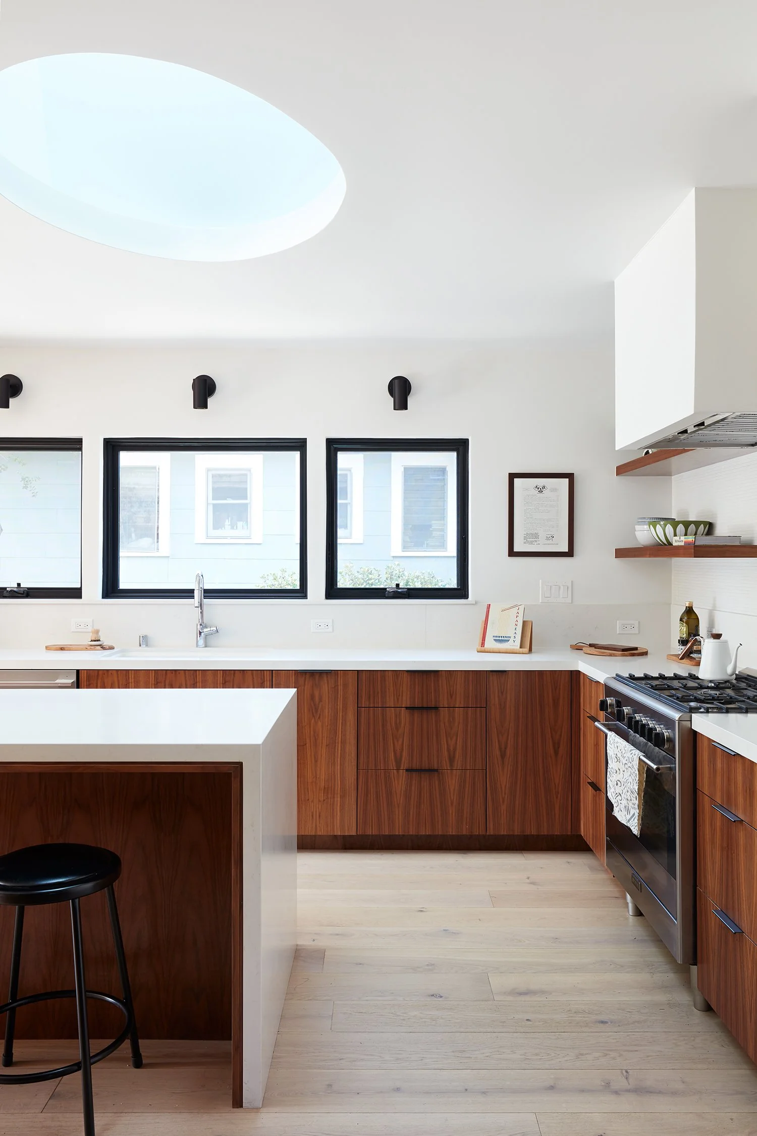 A modern kitchen with white walls, three black-framed windows, and wooden lower cabinets. There is a white countertop, a black stool, and a stainless steel stove with a towel hanging. Open wooden shelves hold bowls and dishes, and a framed document o