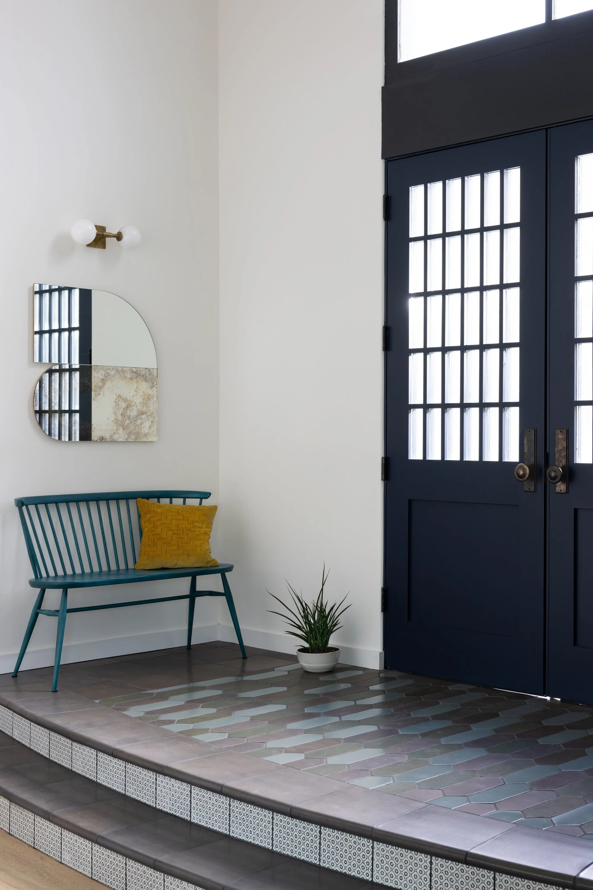 Entryway with dark blue door featuring glass panels, a blue chair with a yellow pillow, a potted plant, and a mirror on a white wall with a light fixture.