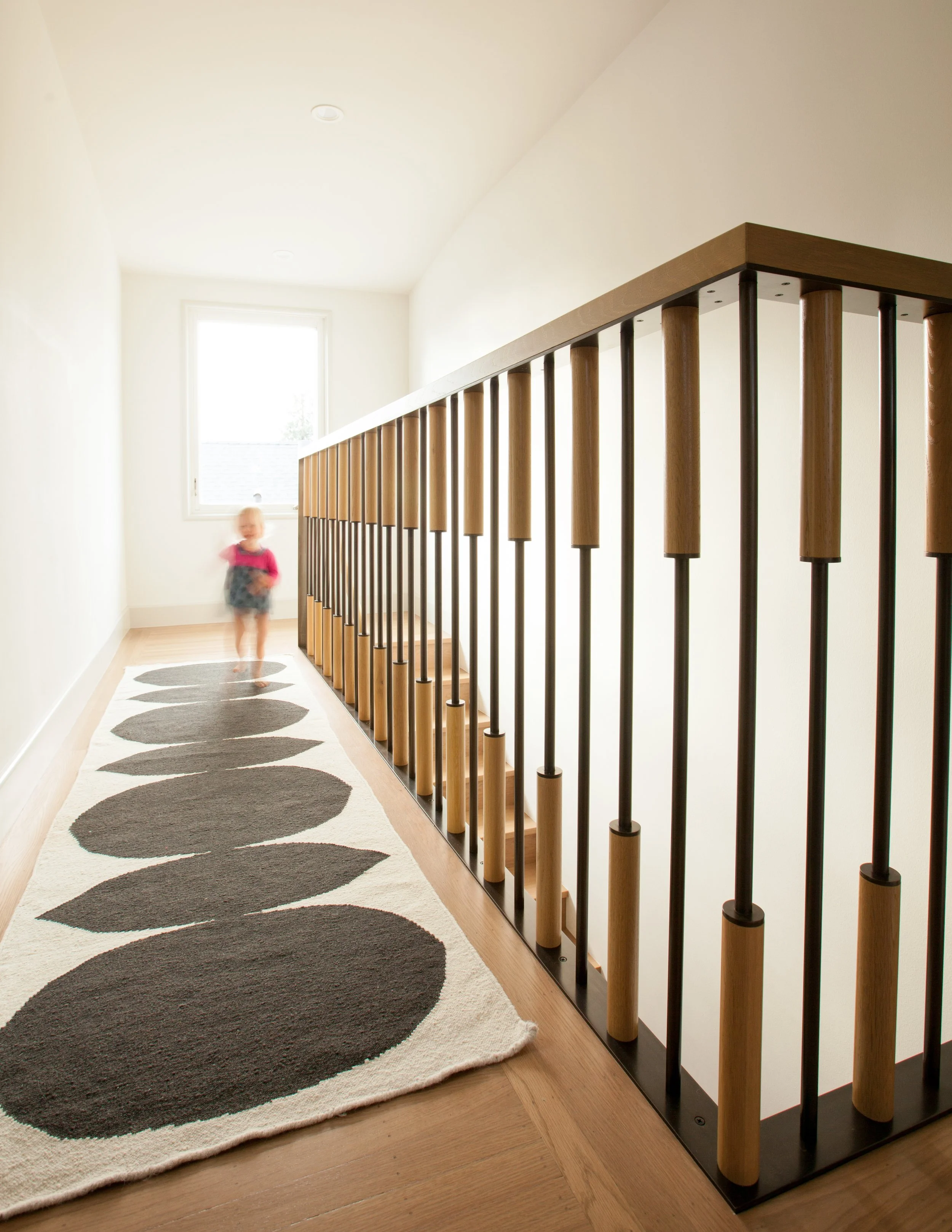 A hallway with a white wall, a large window, a black and white oval patterned rug, and a wooden and metal staircase railing.